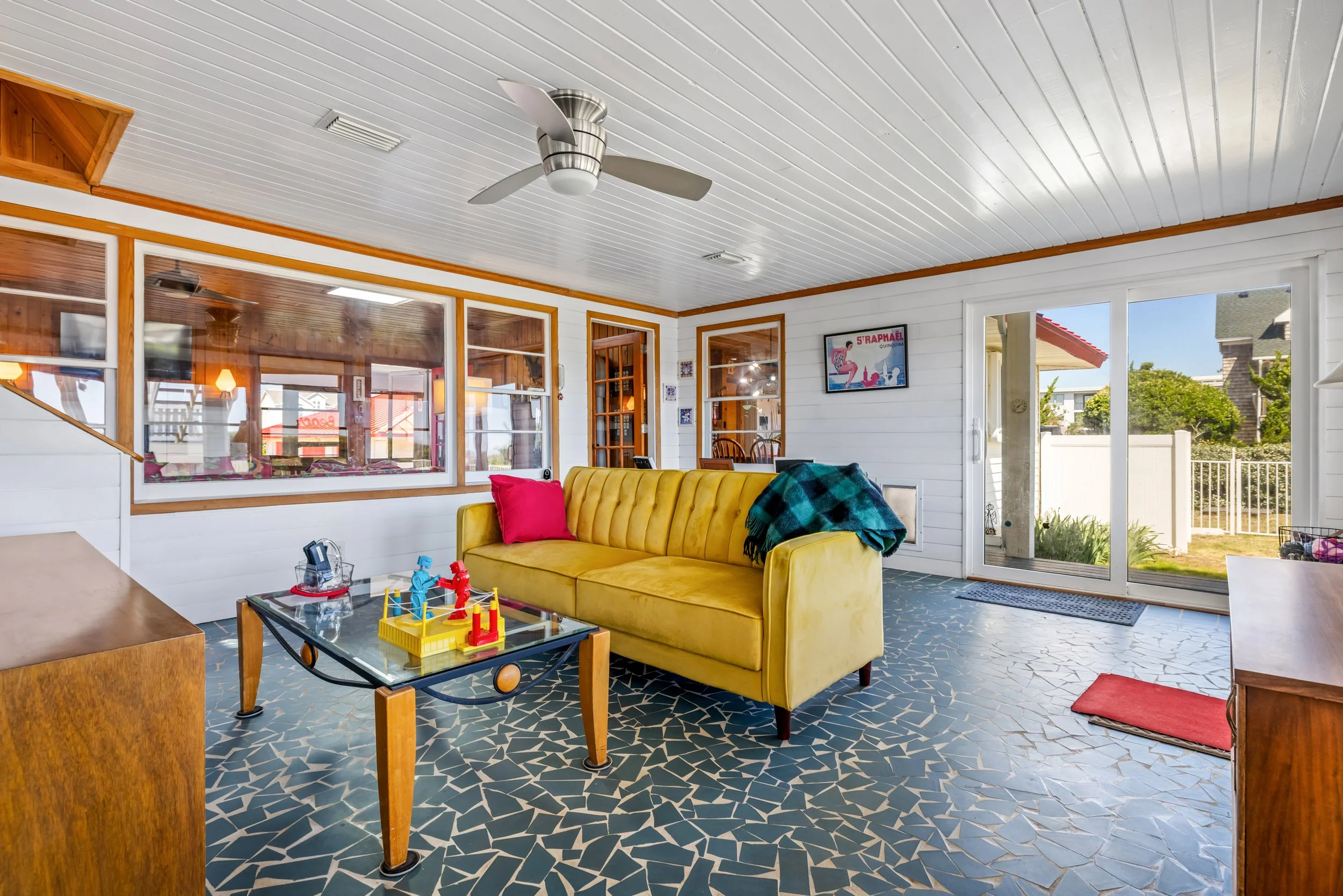 An enclosed patio or sunroom with a white ceiling, a ceiling fan, and large windows and sliding glass door providing outside views. Inside, there is a yellow sofa with colorful cushions, a glass table with a toy game, and a red rug on the floor.