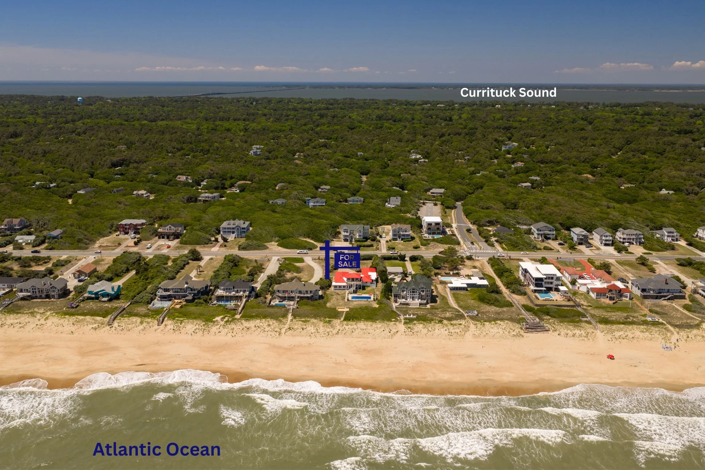 Aerial view of a beachside neighborhood with houses, greenery, and the Atlantic Ocean in the background. Beachfront with sand and ocean waves in the foreground. Label indicating 'Atlantic Ocean' at the bottom and 'Currituck Sound' over the water in t