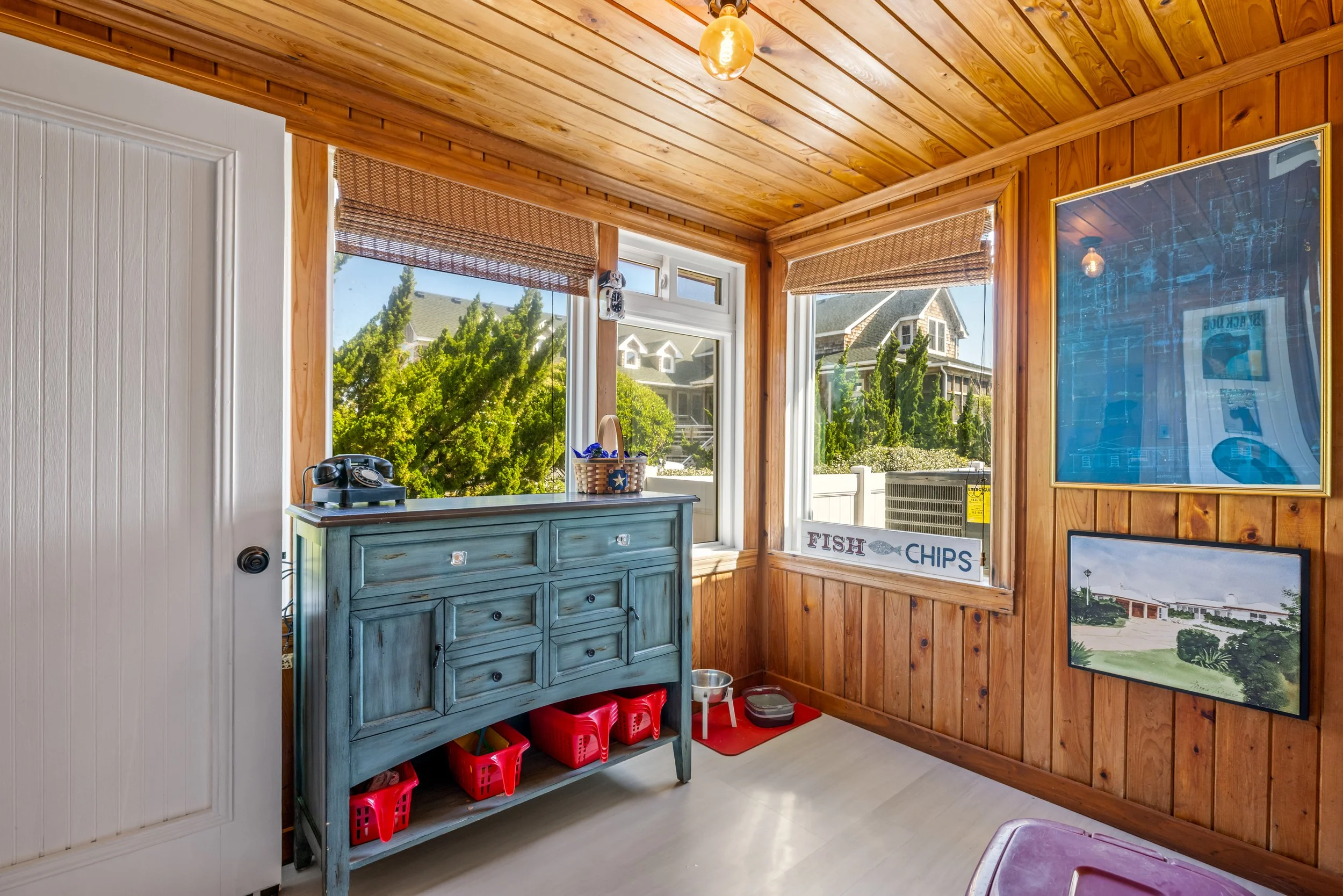 Interior view of a cozy corner in a house with wood-paneled walls and ceiling, large windows showing a sunny neighborhood with trees and houses, a vintage blue chest of drawers, a sign reading 'FISH & CHIPS', and framed pictures.