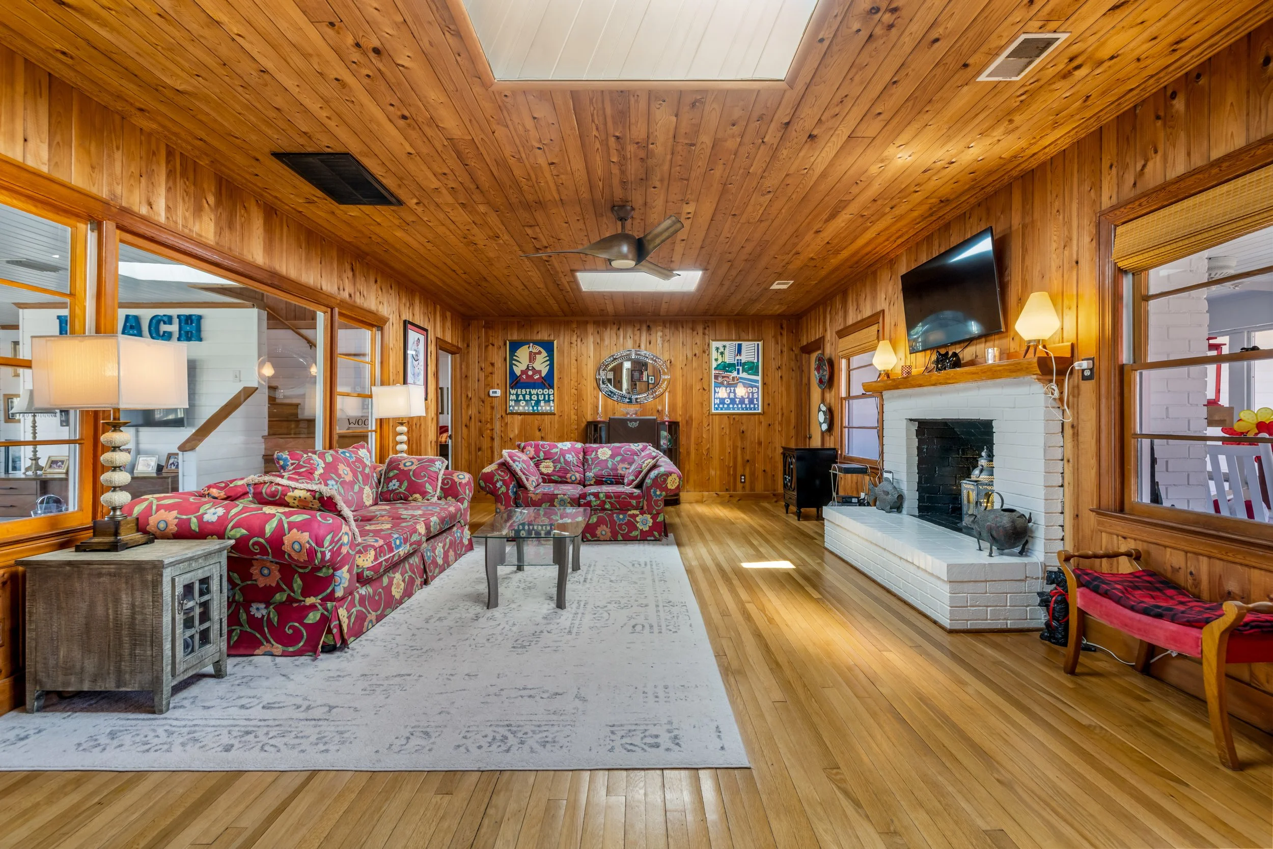 Living room with wood-paneled walls and ceiling, featuring a ceiling fan and skylight. There is a floral patterned sofa, a coffee table, lamps, framed art, a large flat-screen TV, and a white brick fireplace.