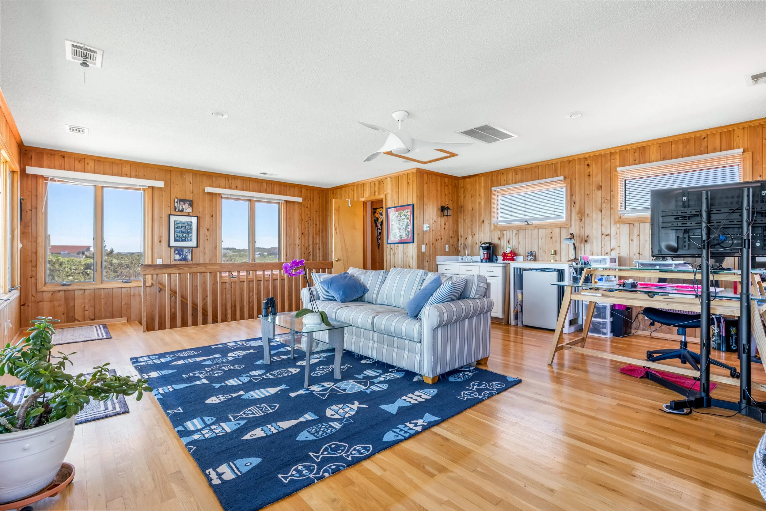 A living room with wooden paneled walls, large windows, a striped sofa, a blue patterned rug, potted plants, and various furniture and decor.