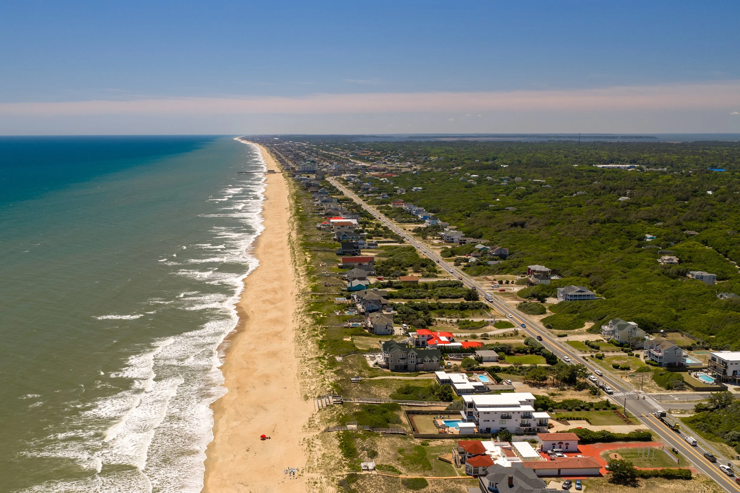 Aerial view of a beach town with houses along the shore, sandy beach, waves, and a road parallel to the coast, with greenery on the right.