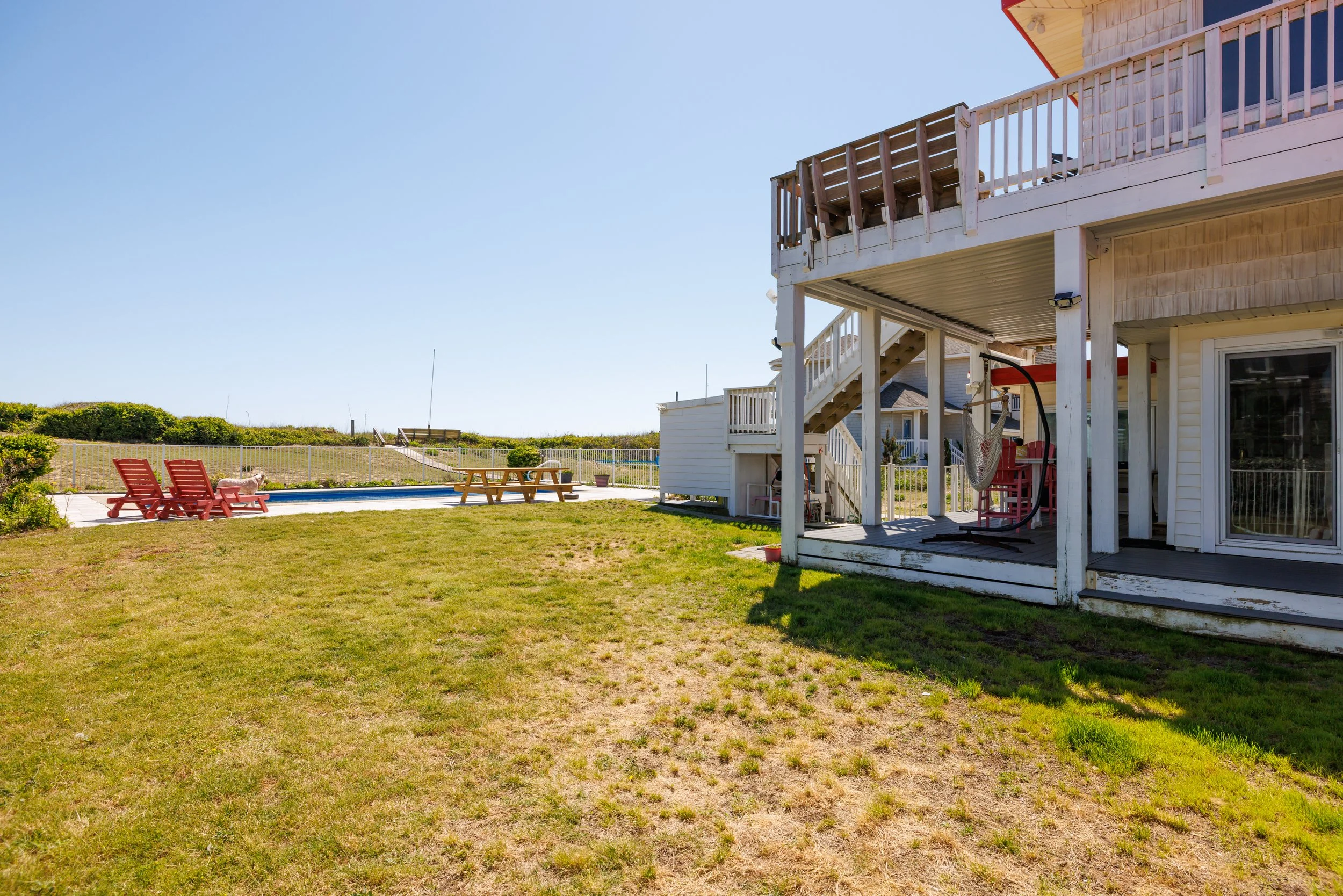 A backyard with a grassy lawn, two red lounge chairs near a swimming pool, a picnic table, and a white house with a balcony and stairs. There is a dog on the lawn and the sky is clear and sunny.
