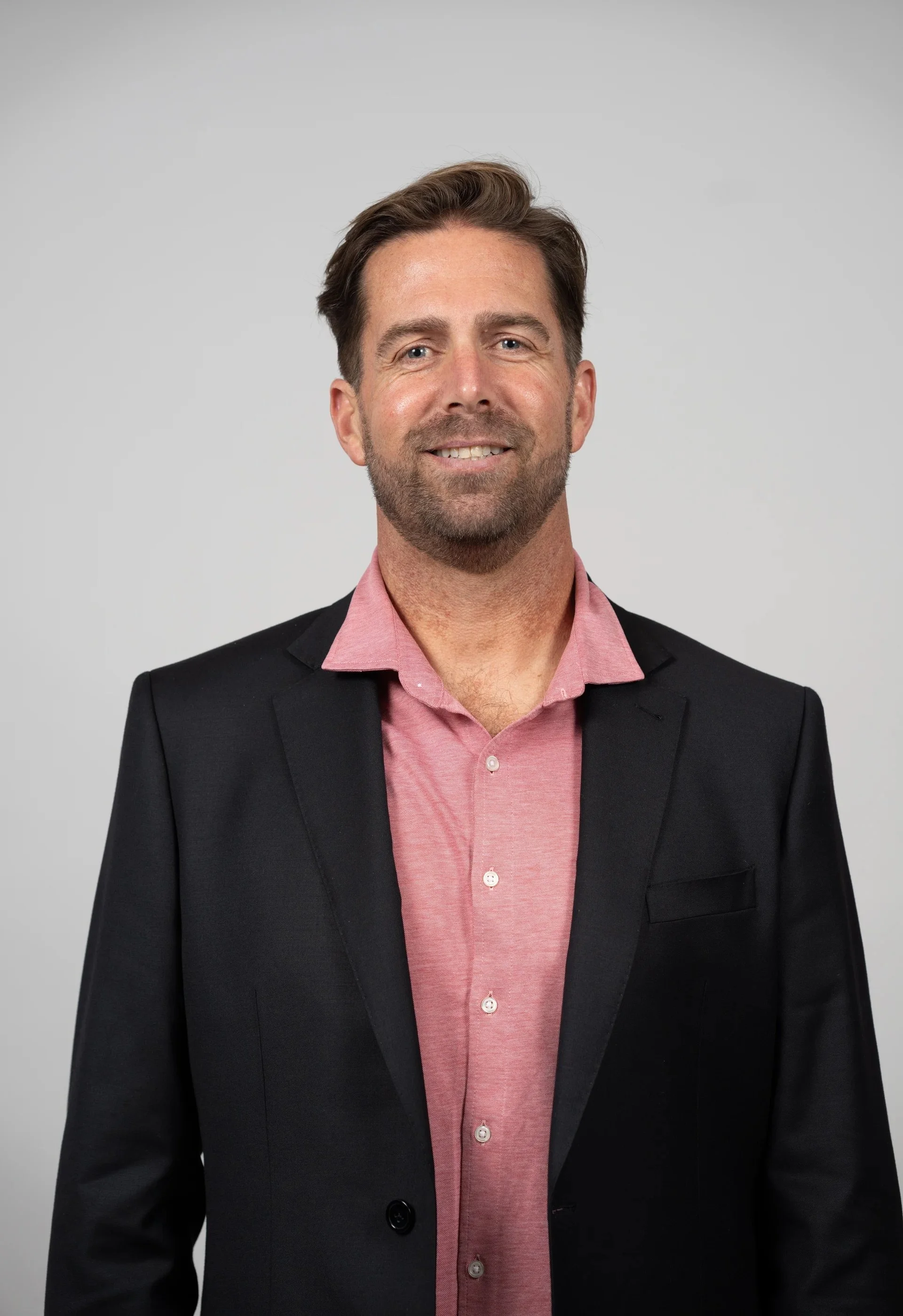 A man with brown hair and a beard, wearing a black blazer and pink shirt, smiling against a plain gray background.