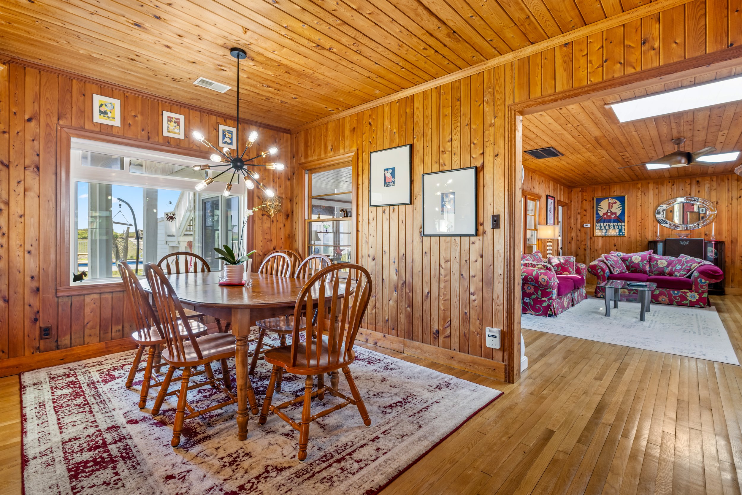 A dining area with a wooden table and chairs, decorated with a potted plant. The room has wooden paneling on the walls and ceiling, with several framed art pieces. In the background, there is a living area with floral sofas, a mirror, and a ceiling f