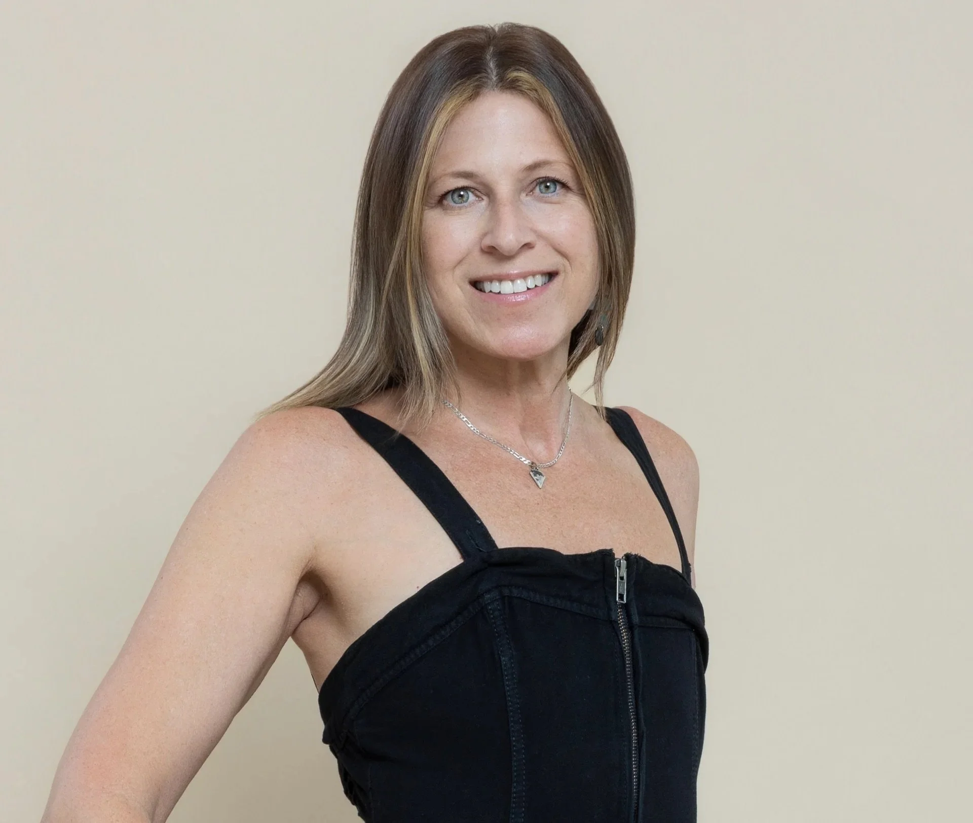 A woman with shoulder length brown hair and blue eyes wearing a black women sleeveless top with zip front and a silver necklace, standing against a plain beige wall.