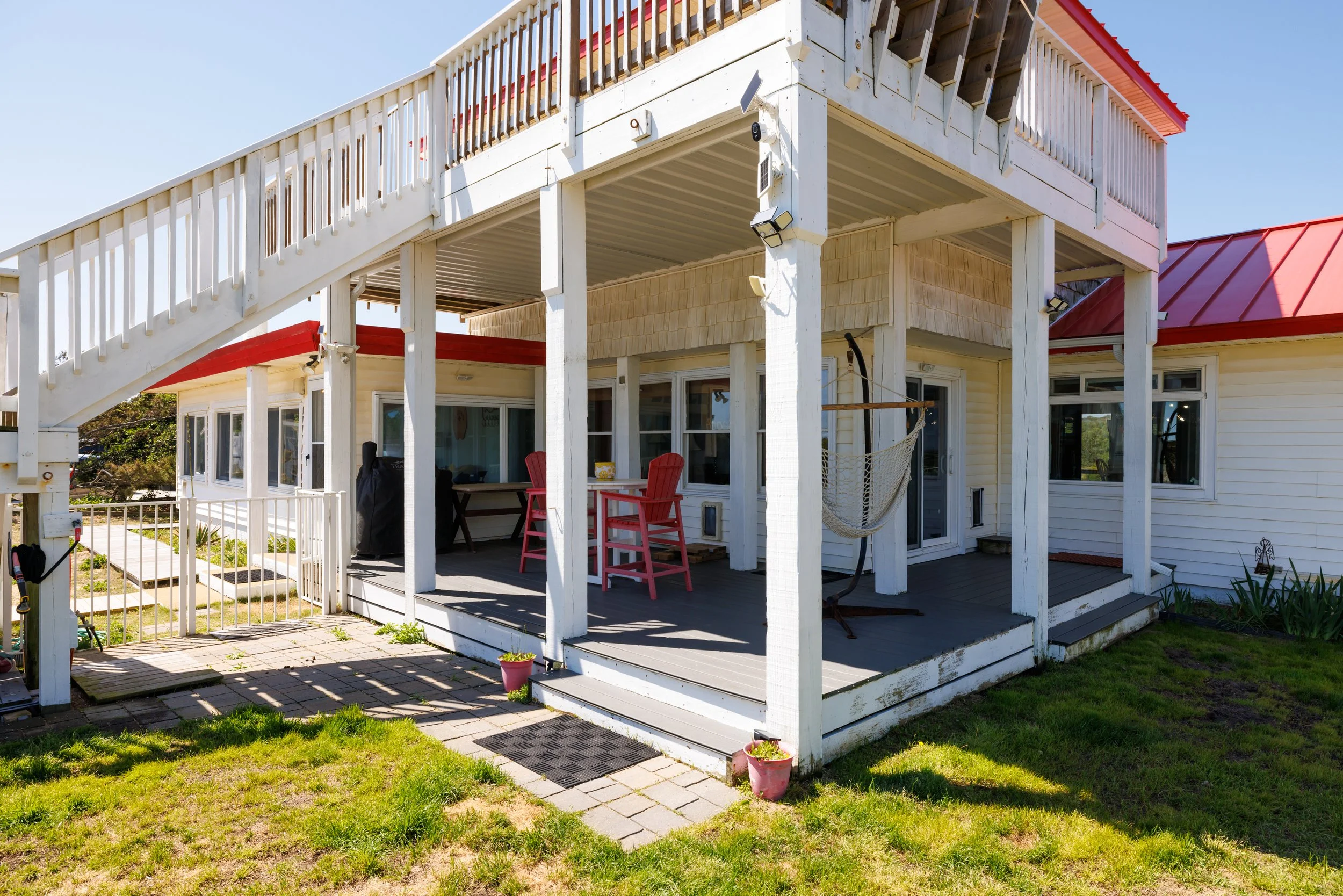 The image shows the back of a two-story house with a large deck. The house has white siding and large windows. There is outdoor furniture, including two pink chairs and a table, on the deck. A staircase with white railings leads from the deck to the 