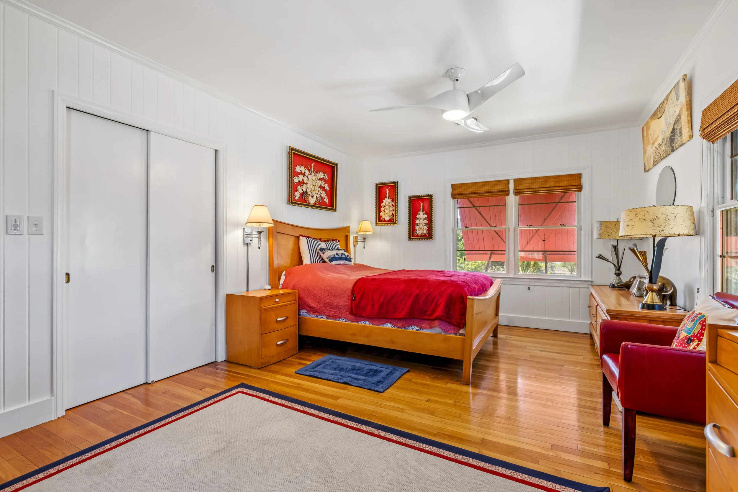 A bright bedroom with a wooden bed frame and a red bedspread, surrounded by nightstands and lamps, with framed floral artwork on the white-paneled walls and two windows with wooden blinds.