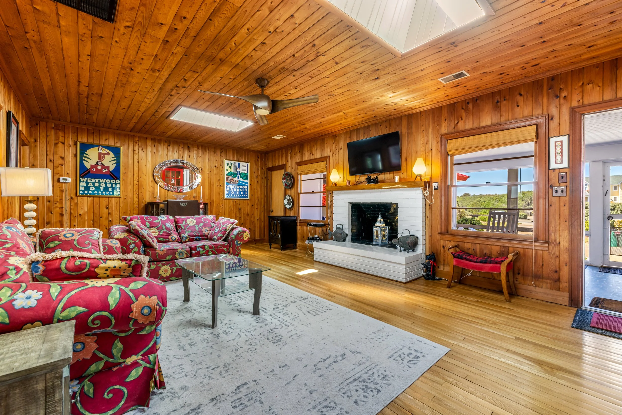 A living room with wood-paneled ceiling and walls, a fireplace with white brick, a flat-screen TV above it, colorful floral-patterned sofas, windows with blinds, and decorative artwork on the walls.