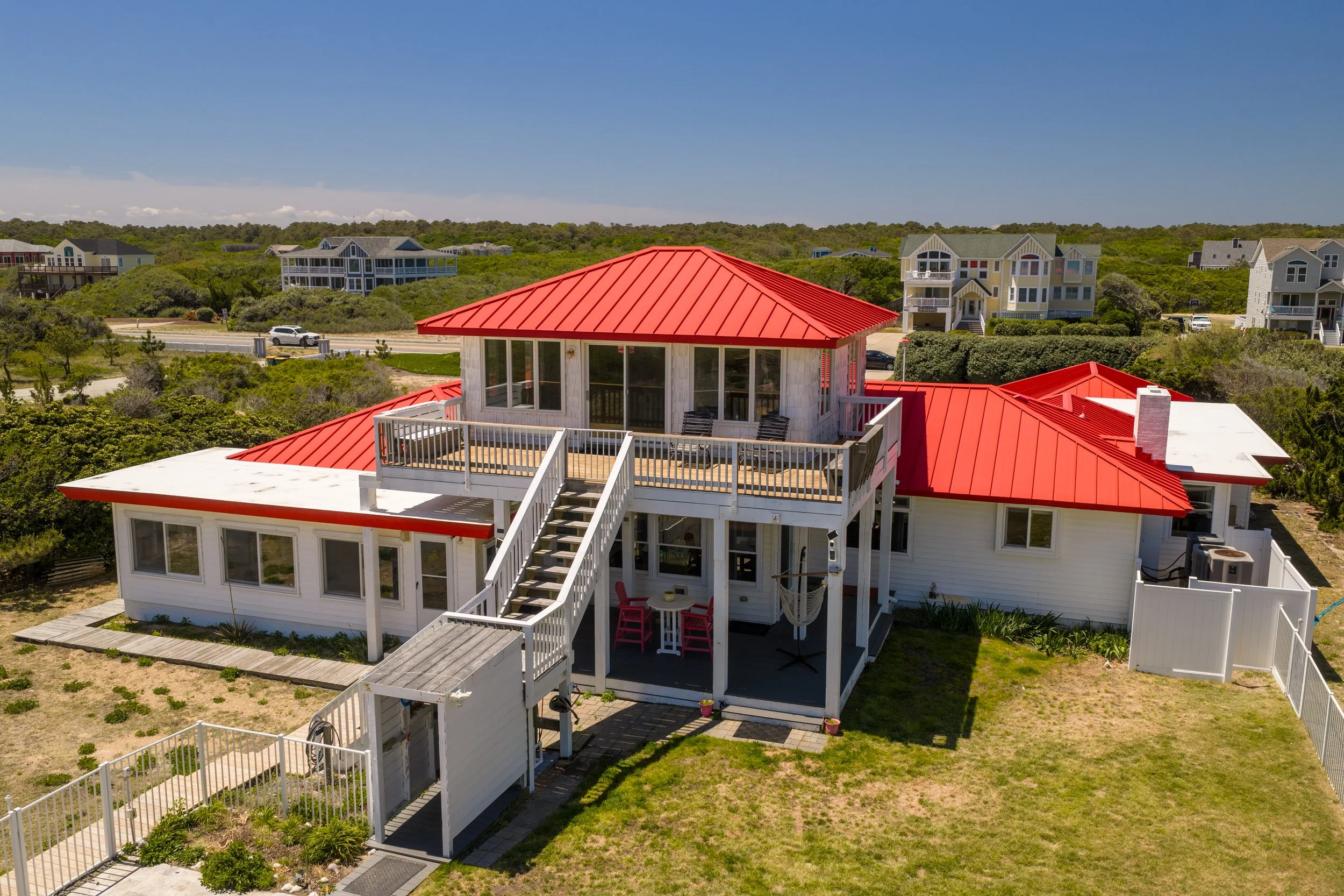 A house with a red metal roof, multiple decks, and large windows, set in a neighborhood with other houses, bushes, and trees under a blue sky.