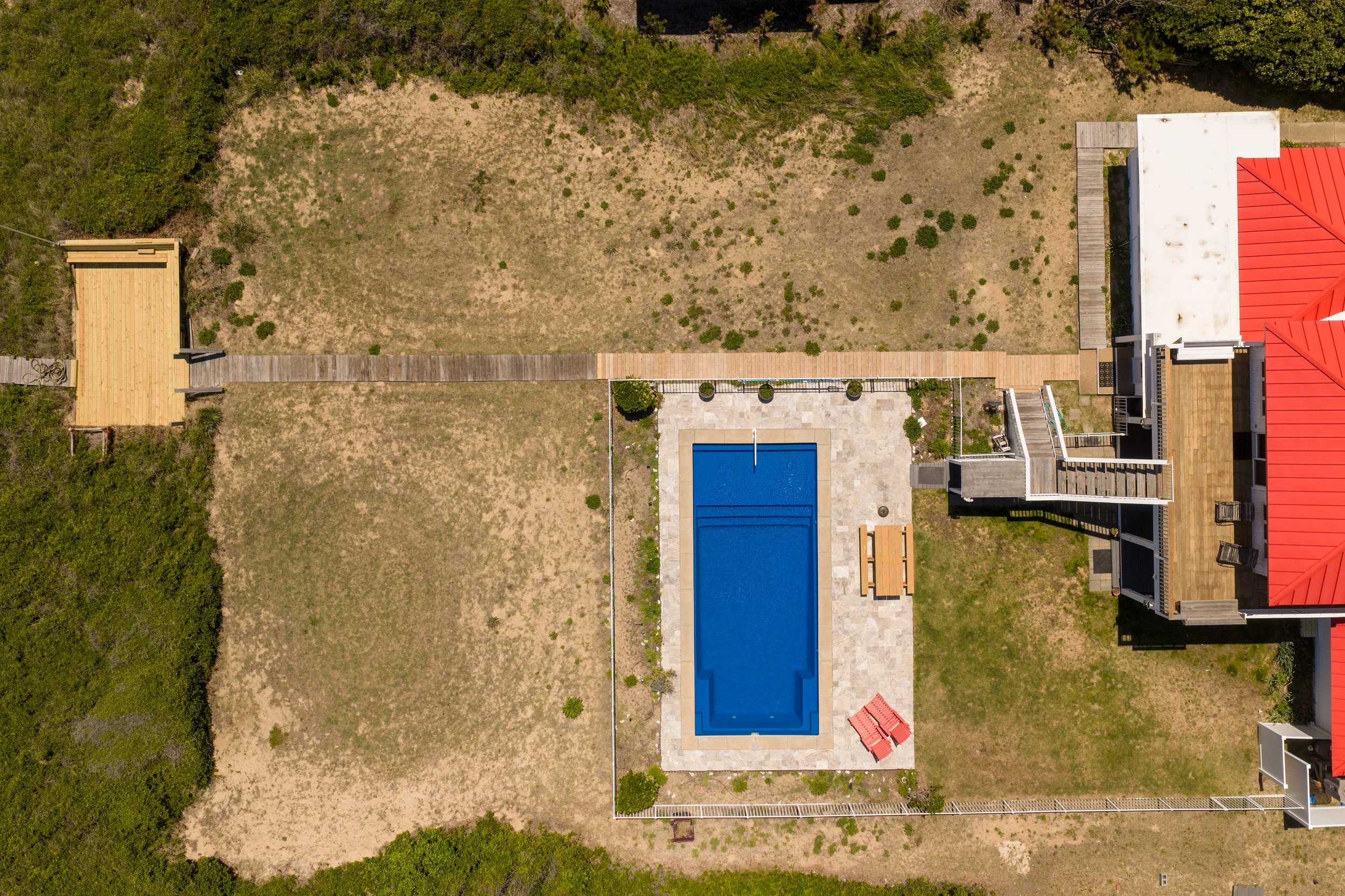An aerial view of a backyard with a rectangular swimming pool, a patio area with two red umbrellas, a wooden deck, a brightly colored house with a red roof, and a grassy lawn around it.