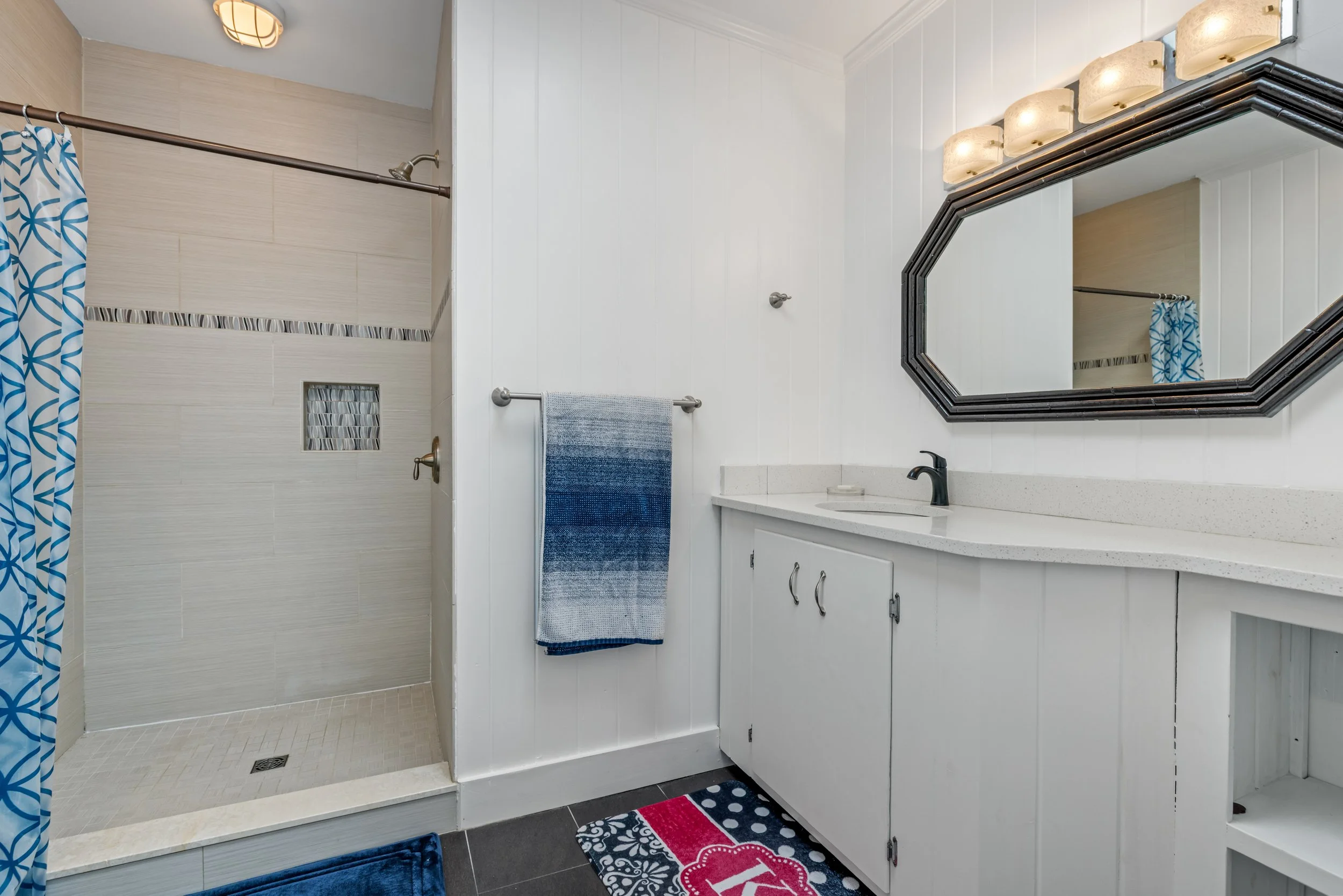 A bathroom with a walk-in shower on the left, featuring a blue patterned shower curtain. The shower has beige tiles and a small window with decorative tile trim. On the right, there's a white vanity with a speckled countertop, a black faucet, a large
