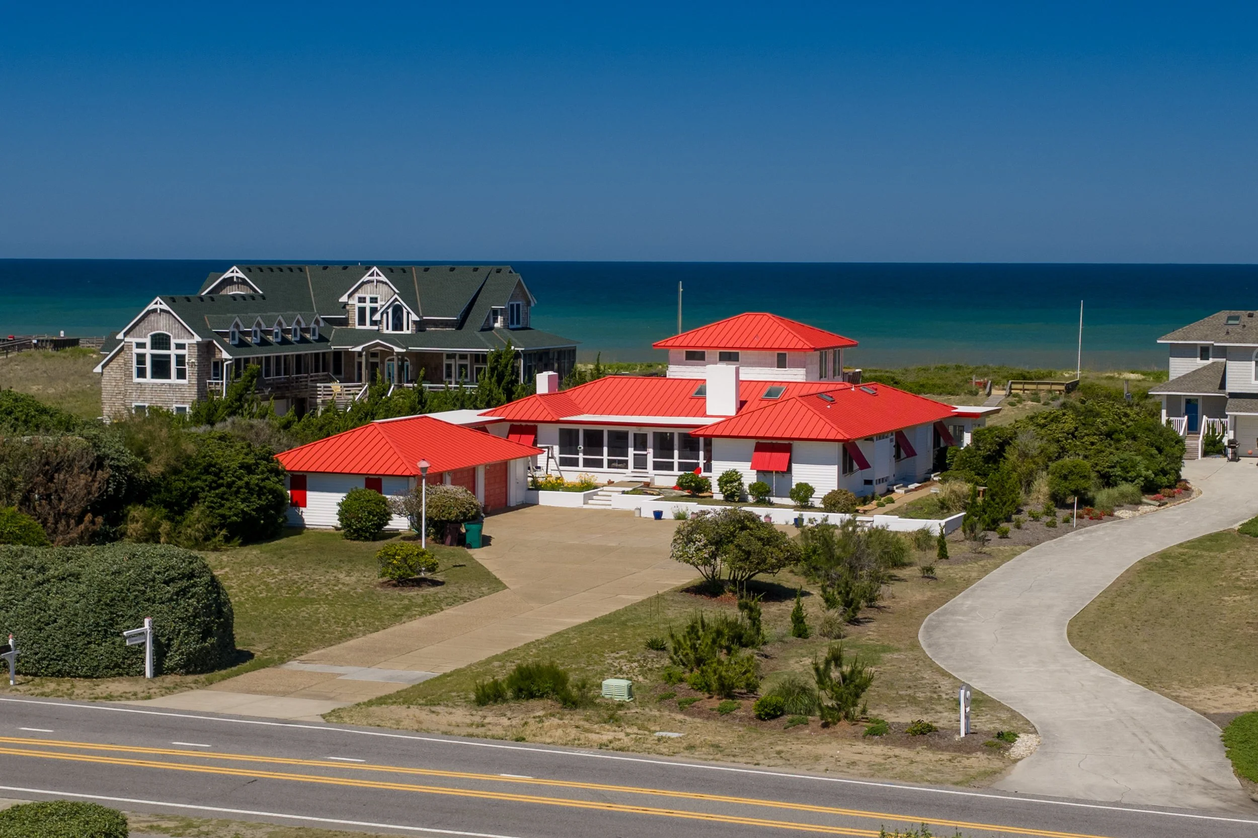 A coastal neighborhood with large houses featuring red and gray roofs near the ocean under a clear blue sky. A winding path and sidewalk are visible in the foreground.