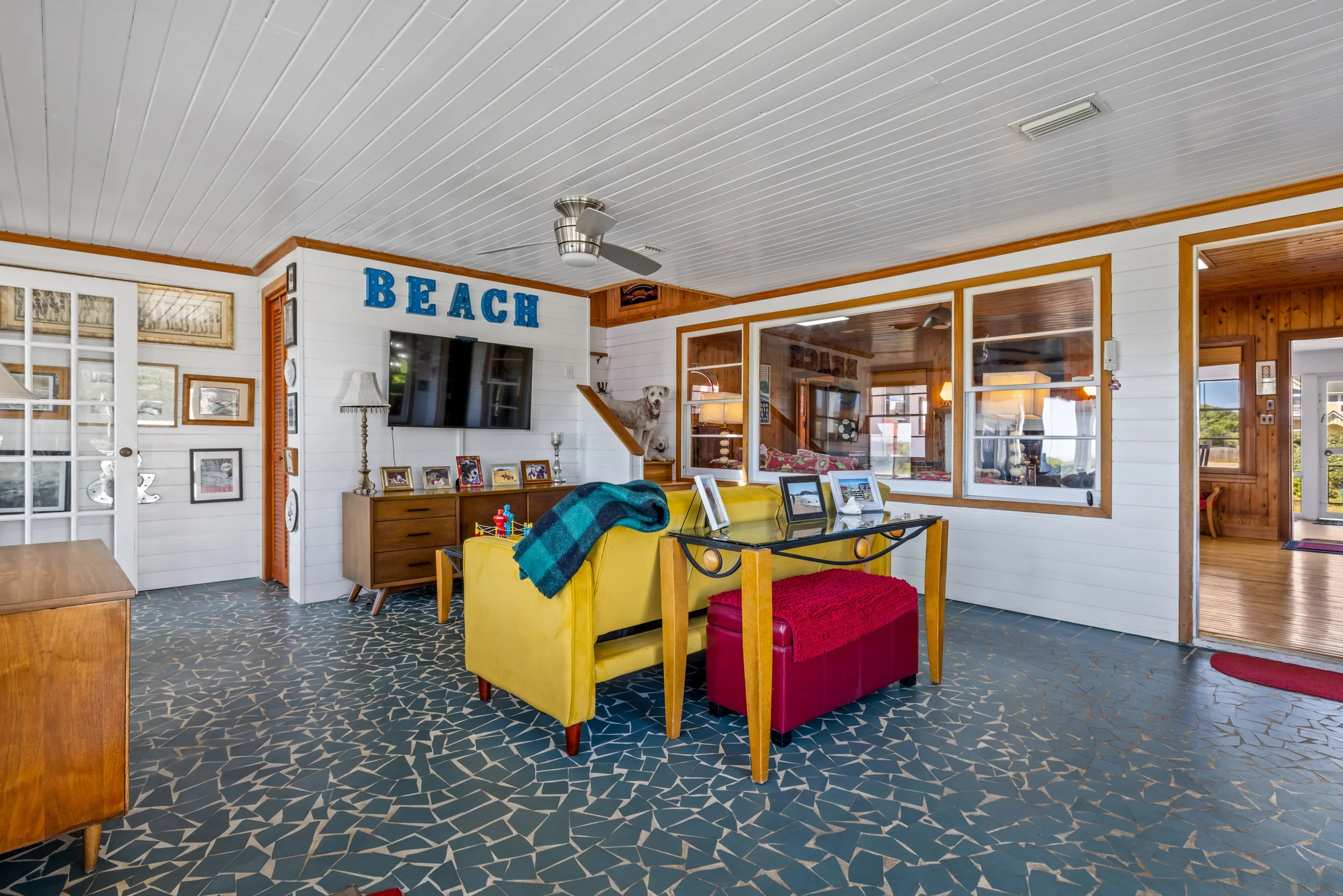 View of a living room with white paneled walls, a patterned blue floor, and mid-century furniture. A yellow sofa with a blanket, a red ottoman, and a wooden sideboard with framed photos and decorative items are visible. A large flat-screen TV is moun