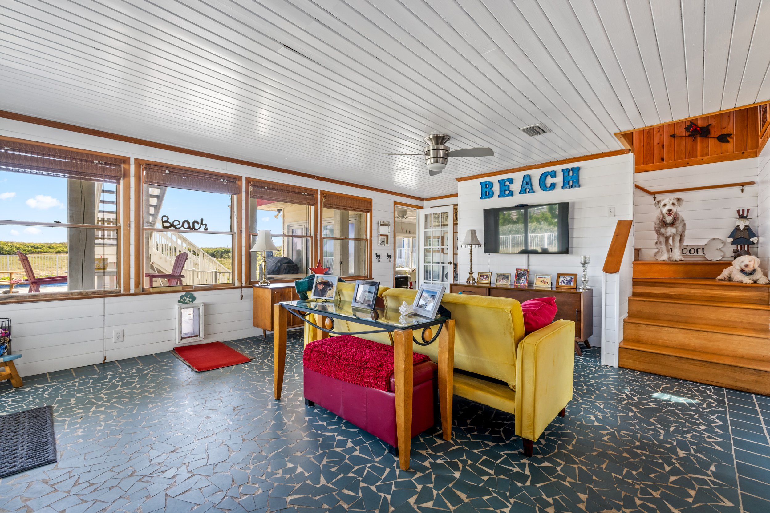 A cozy living room with large windows showing a beach view, decorated with colorful furniture, framed photos, and beach-themed decor including a large sign that says "BEACH" and plush dogs on a staircase