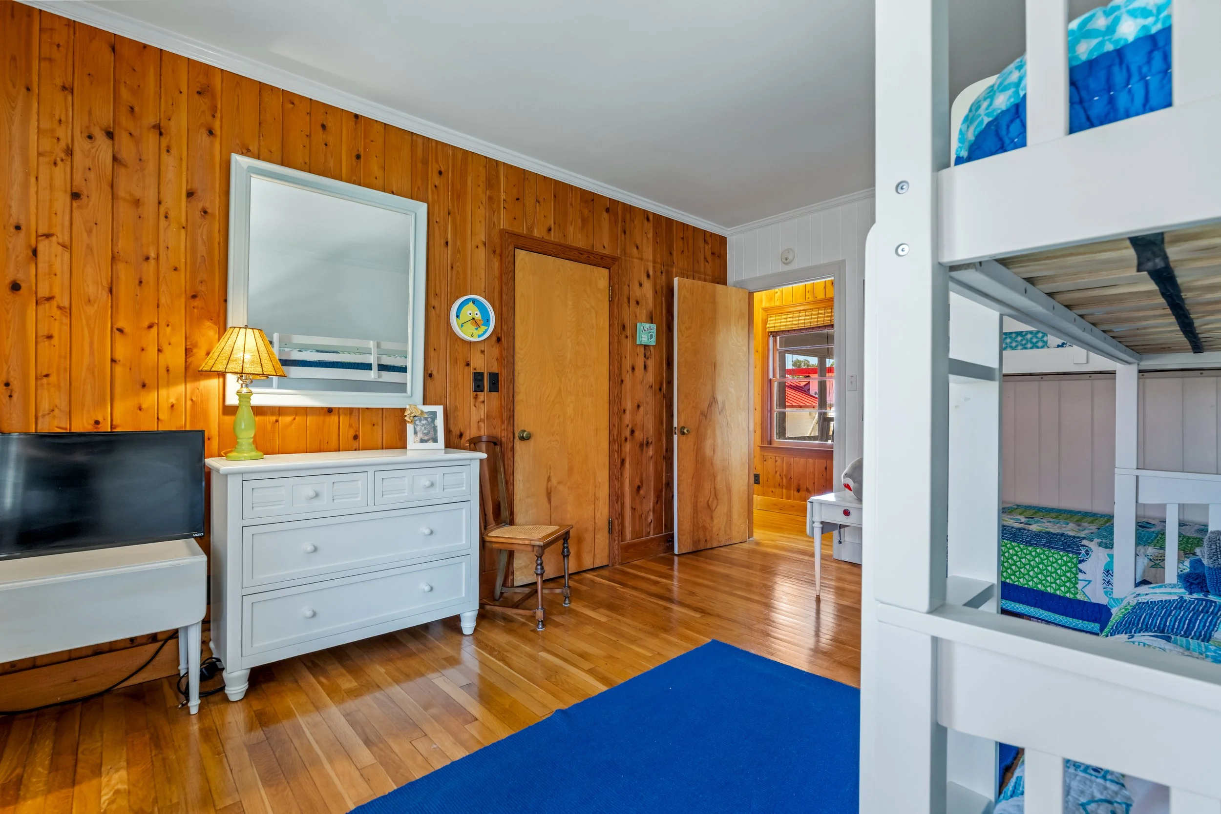 Bedroom with oak wood panel walls, a white dresser with a yellow lamp, a framed photo, and a mirror. There is natural light from a window, a bunk bed with blue bedding, and a blue rug on the hardwood floor.