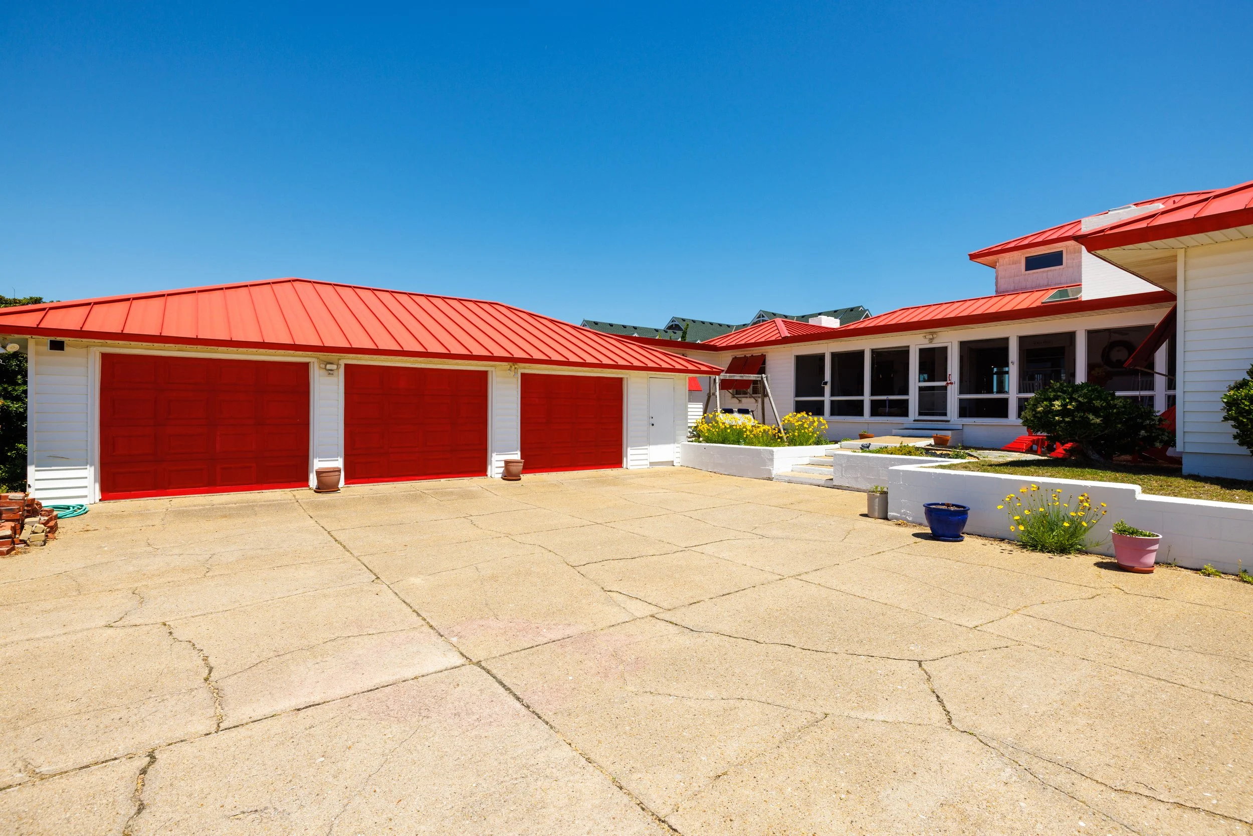 A house with a red roof and matching garage doors, a driveway with a crack, and potted plants along the sidewalk. There is a blue sky above and some neighboring houses in the background.