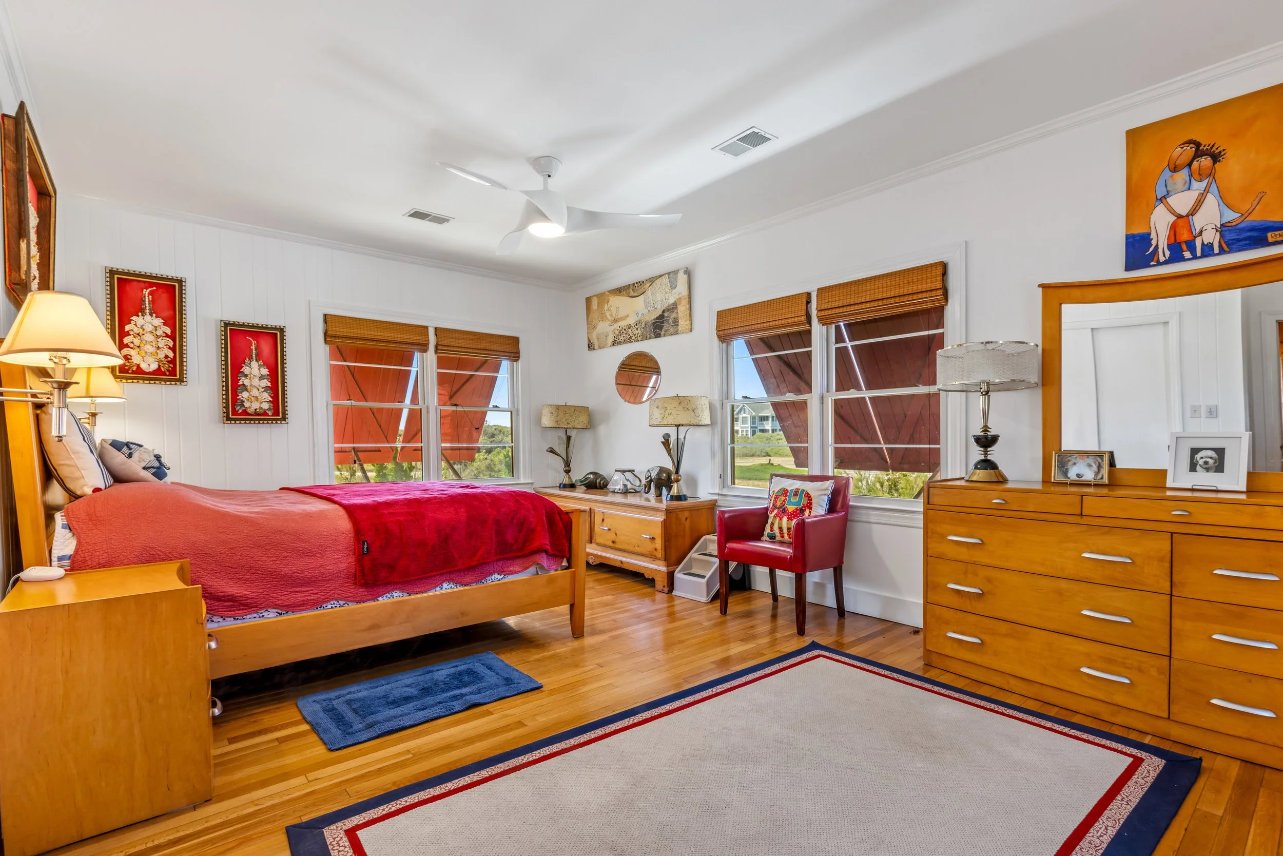 A cozy bedroom with a wooden bed frame, red bedding, and multiple pillows, illuminated by lamps and natural light from windows with wooden shades. The room features wall art, a dresser with framed photos, and a colorful area rug.