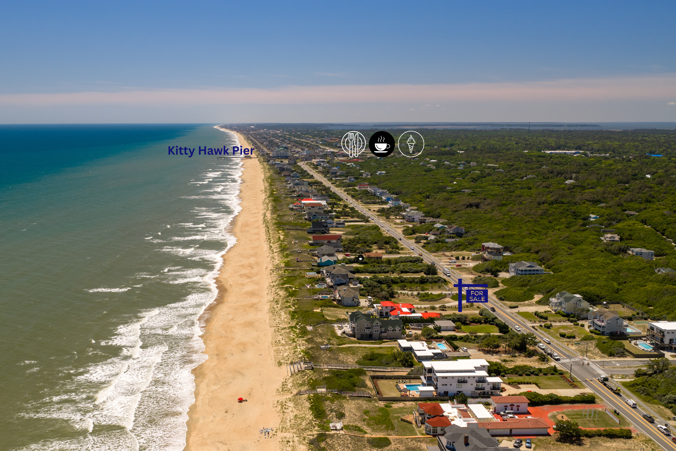 Aerial view of Kitty Hawk Pier along the beach with houses, roads, and green vegetation nearby under a partly cloudy sky.