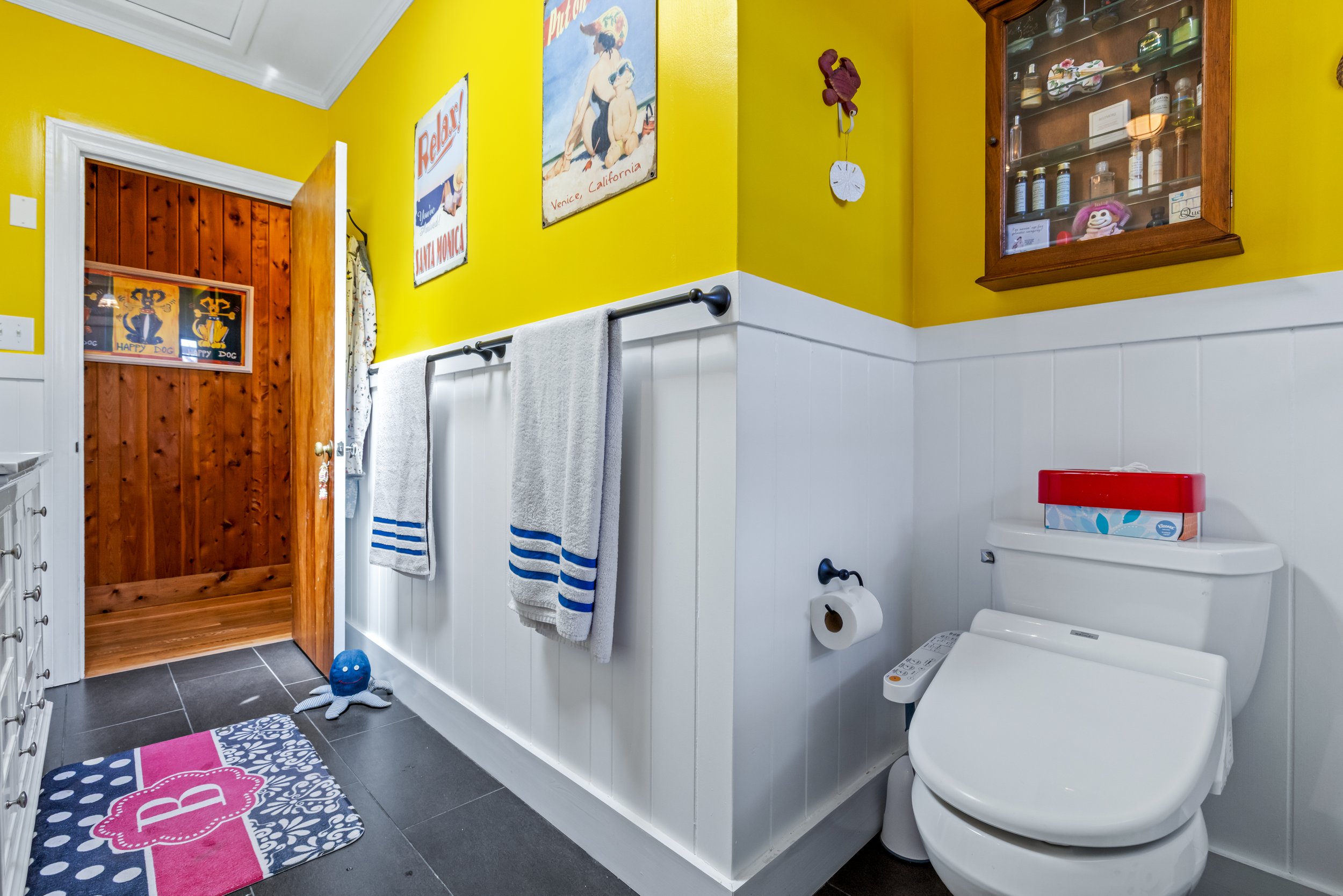 A brightly decorated bathroom with yellow walls and white beadboard wainscoting. There are towels hanging on black rods, artwork on the walls, and a wooden cabinet with glass doors displaying toiletries and a plush toy. A toilet with a box of tissues