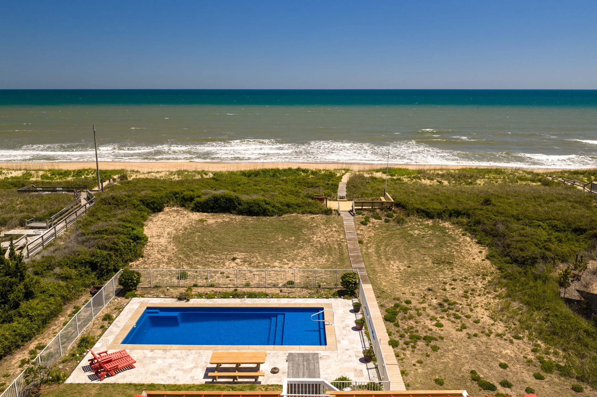 View of a backyard with a swimming pool, patio area with seating, surrounded by greenery, leading to a sandy beach and the ocean in the background under a blue sky.