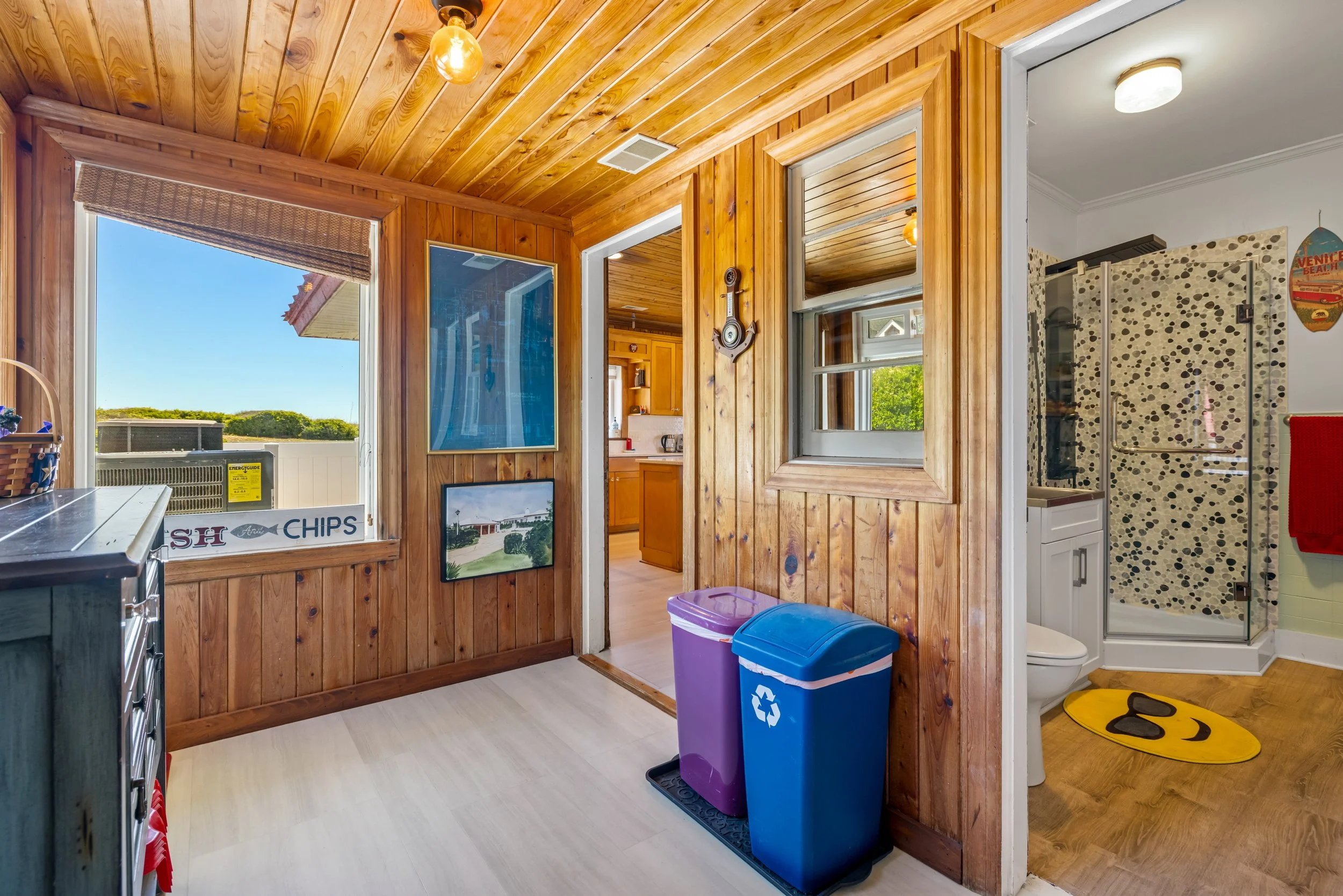 A cozy kitchen with wooden panel walls and ceiling, showing a view into a bathroom with a shower, a window, and a white toilet, with two recycling bins in the foreground.