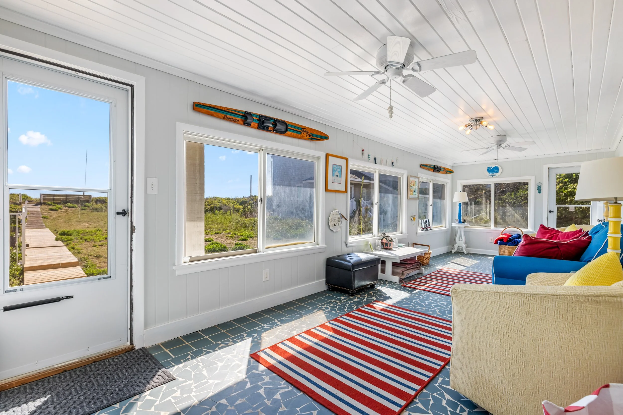 Interior view of a bright living room with white wooden walls and ceiling, multiple large windows, a ceiling fan, a colorful striped rug, a sofa with red and yellow cushions, and beach-themed decorations.