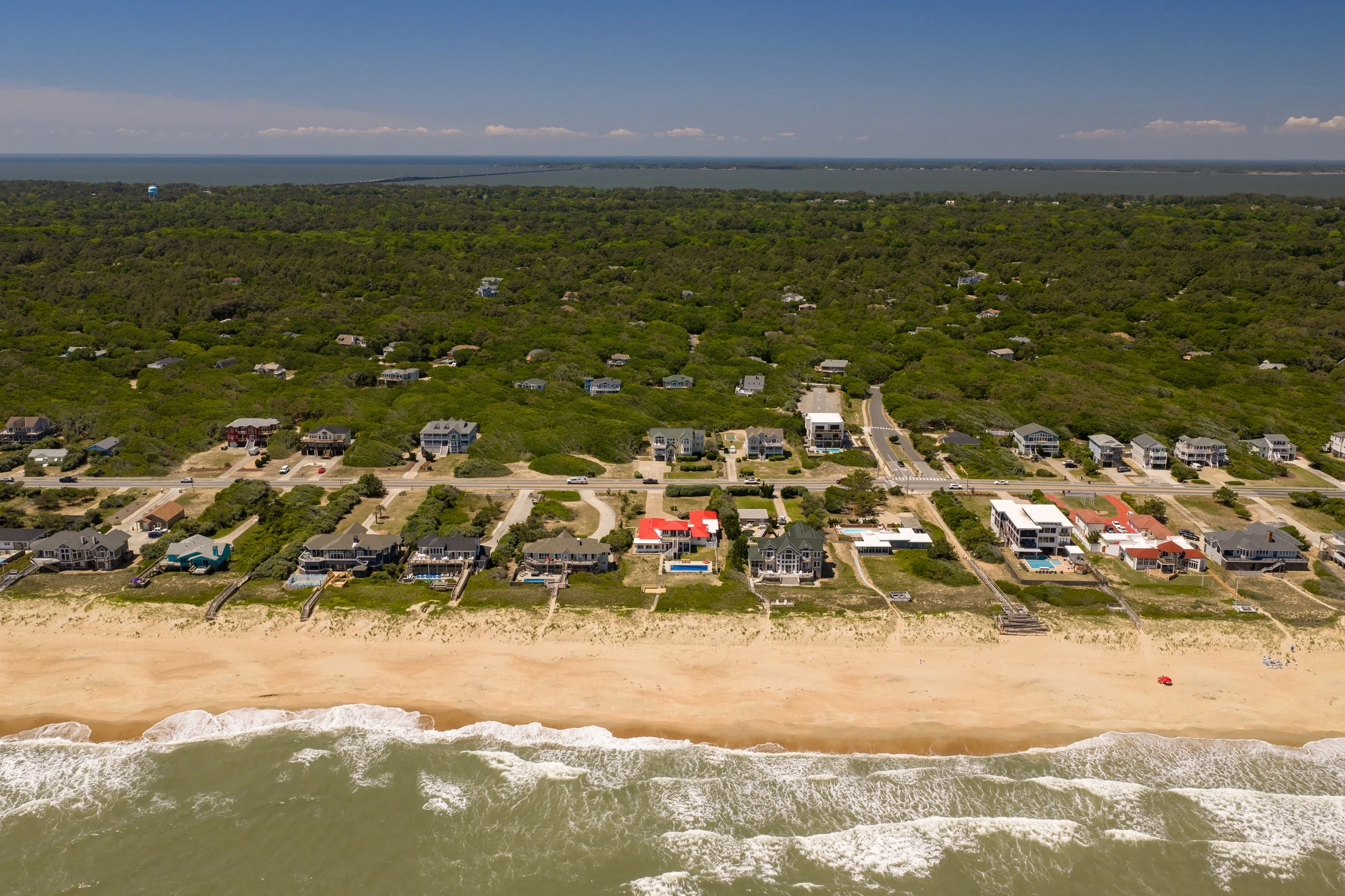 Aerial view of a beach with waves hitting the shore, residential houses, lush green trees, and a river in the background under a blue sky with scattered clouds.