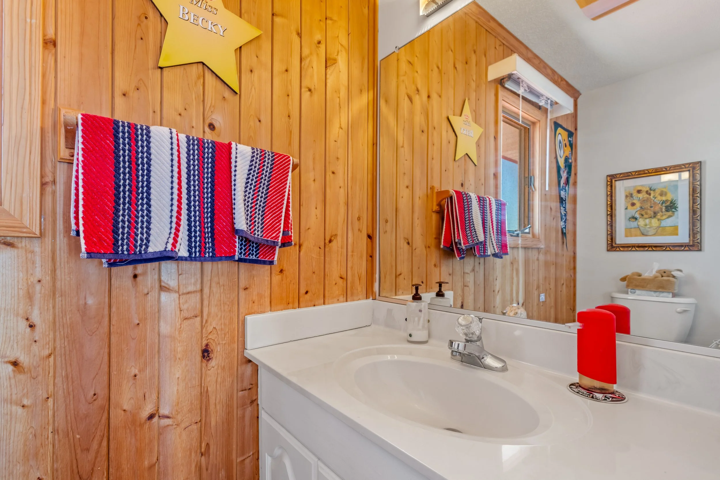 A bathroom with wooden paneled walls, a white sink, and a mirror. Two red, white, and blue striped towels hang on hooks on the wall. There are yellow star decorations and a framed picture of sunflowers on the wall. A window with blinds and a faucet w