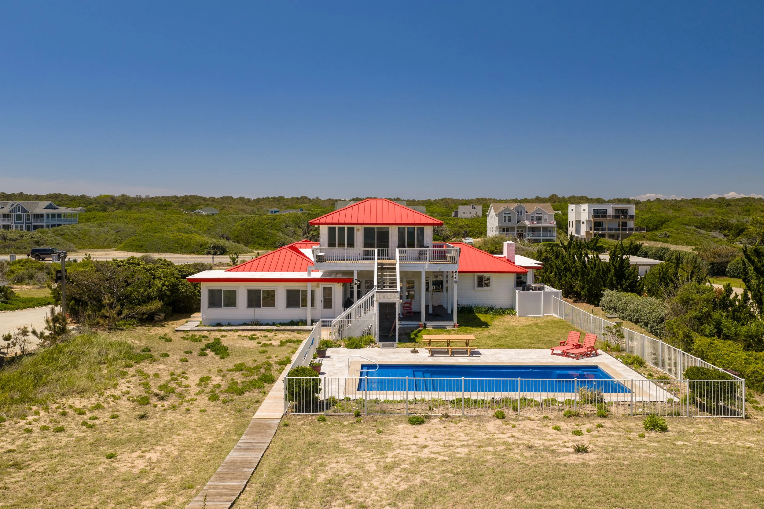 A two-story house with a red metal roof, a backyard pool, and a surrounding white fence. There is a wooden walkway leading to the house, and an outdoor seating area with red chairs near the pool. The background features multiple houses and green hill