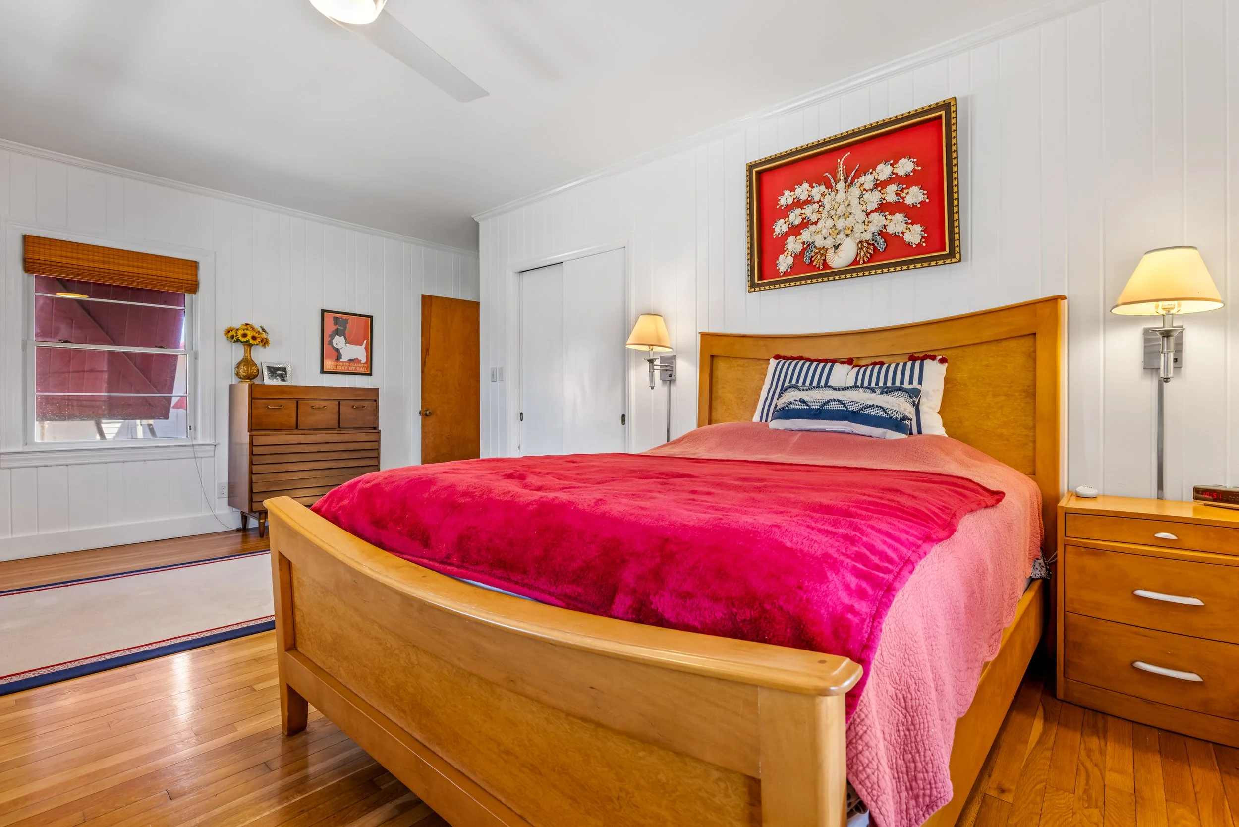 A cozy bedroom with a wooden bed frame and a red bedspread. The bed has embroidered pillows, and there are two wall-mounted lamps on either side. The walls are white with vertical paneling, and there is a window with wooden blinds on the left. A wood