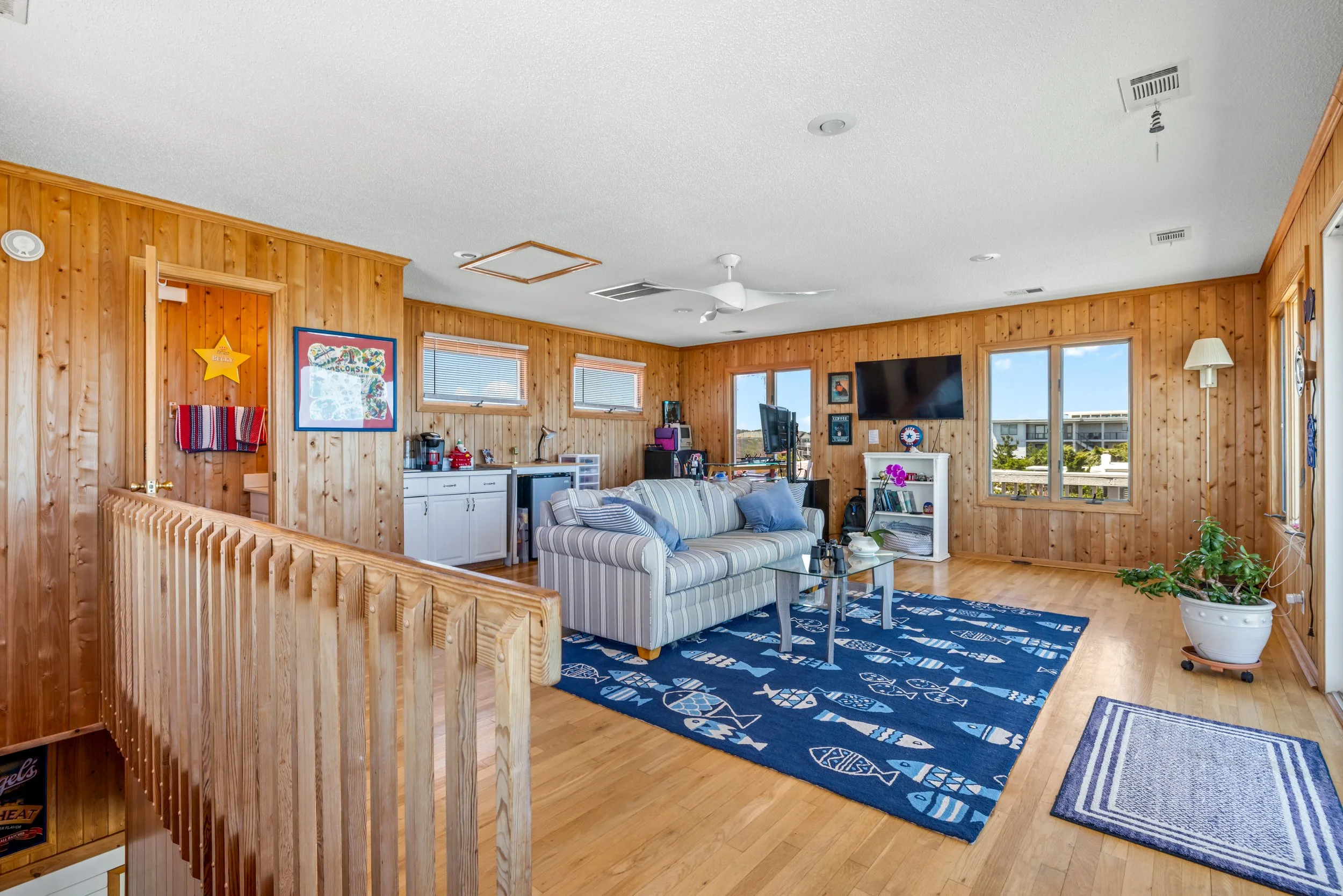 Living room with wood-paneled walls, blue and white patterned rugs, striped sofa, potted plant, large window, wall-mounted TV, and various decor items.