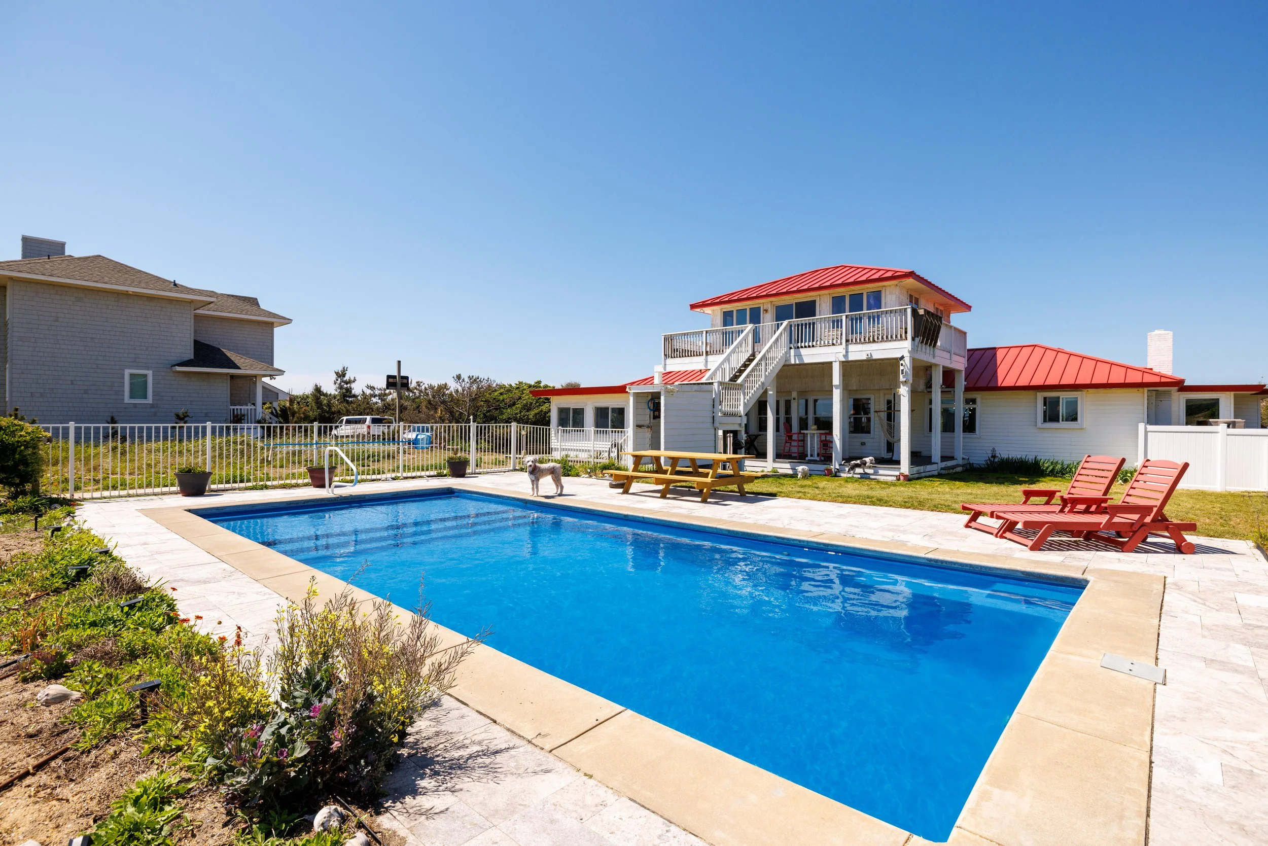 A backyard swimming pool with blue water surrounded by a concrete deck and a white picket fence. Behind the pool, there is a two-story white house with a red roof and a small deck. To the right of the house, there are red lounge chairs on the grass. 