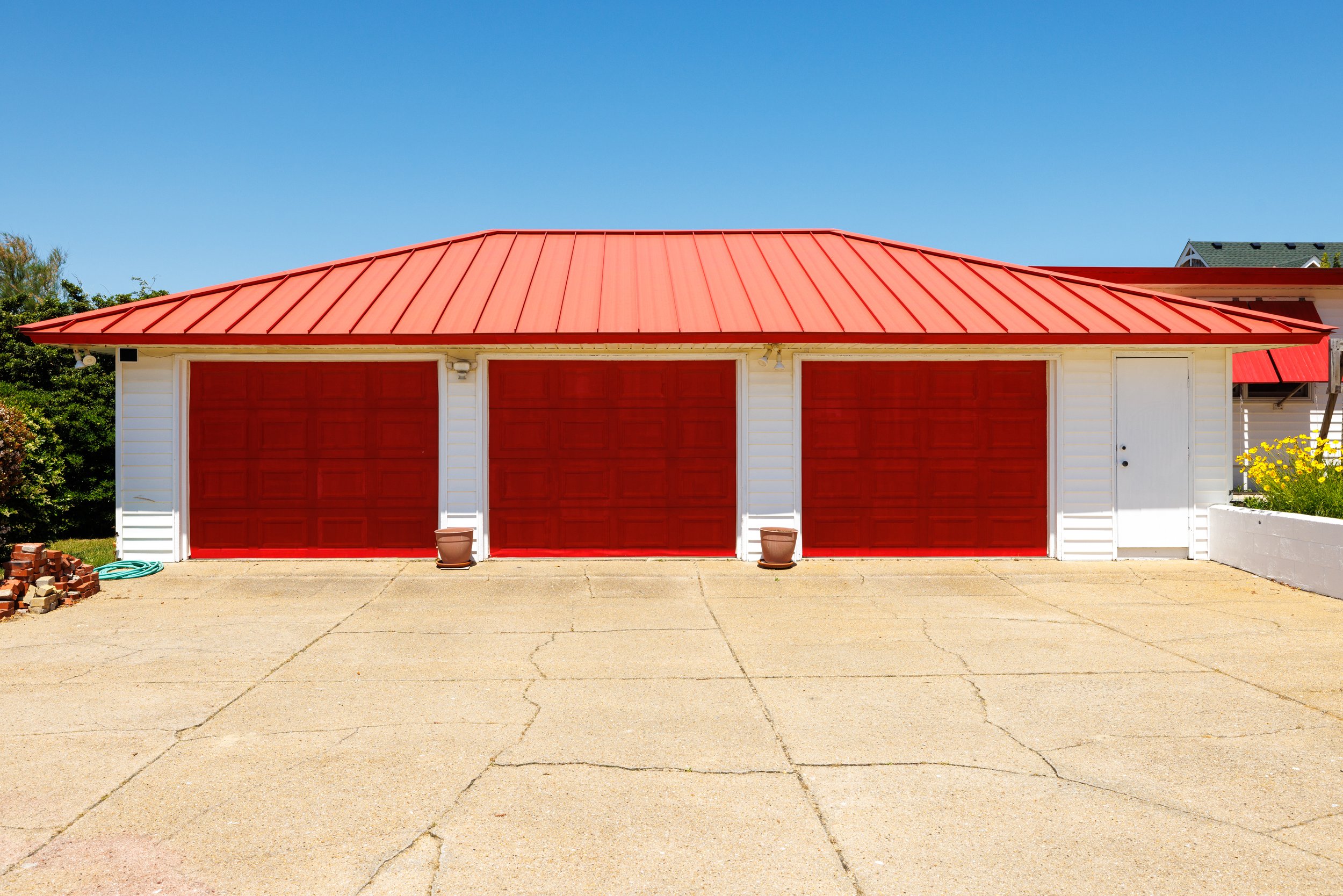 A house with a red metal roof, white siding, and red garage doors, with a concrete driveway in the foreground and some bushes and flowers on the sides.