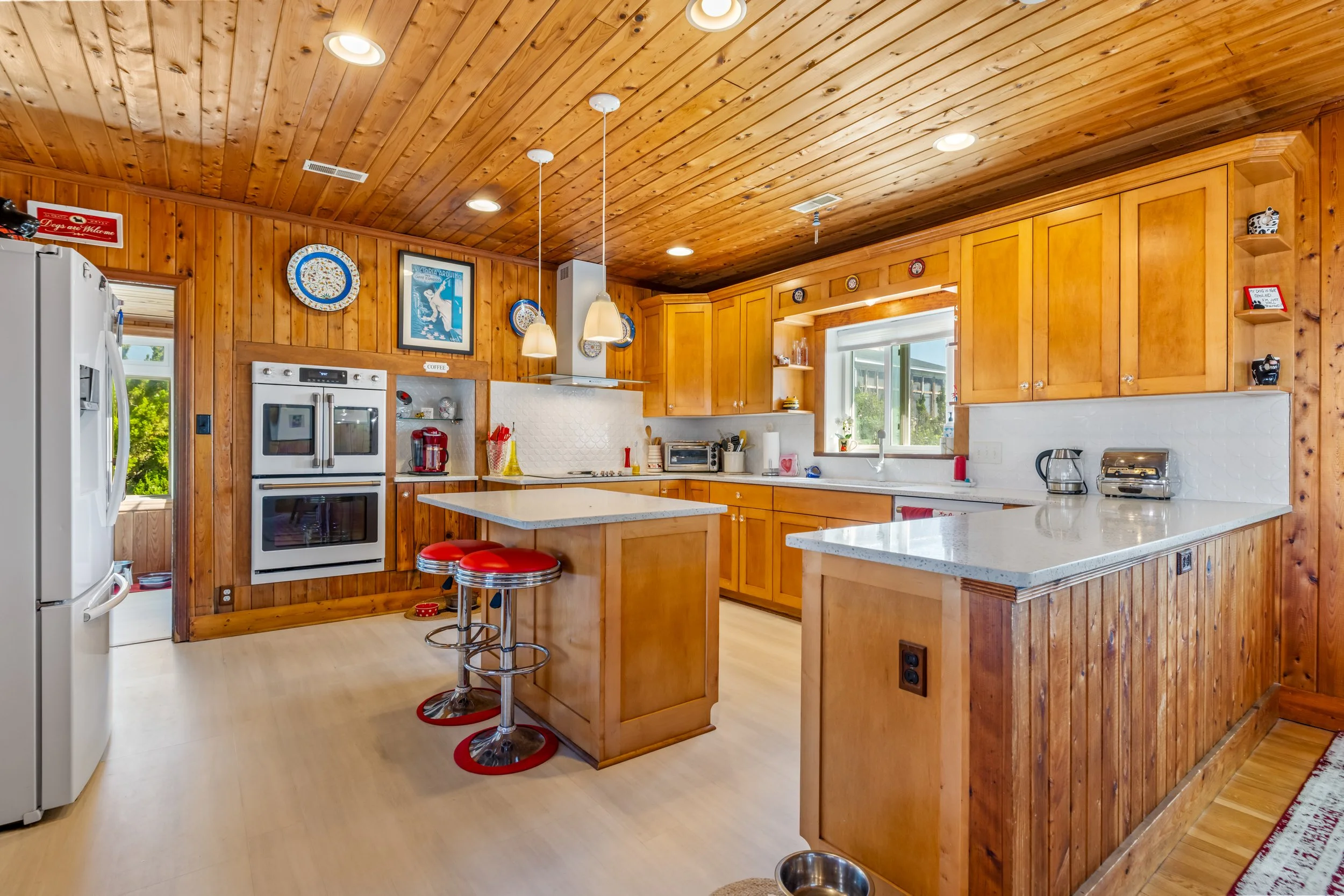 A kitchen with wooden cabinets, a white countertop, and a wooden ceiling. There are kitchen appliances including a refrigerator, oven, microwave, coffee maker, and toaster. Two red stools are at a small island, and decorative plates and framed art ha