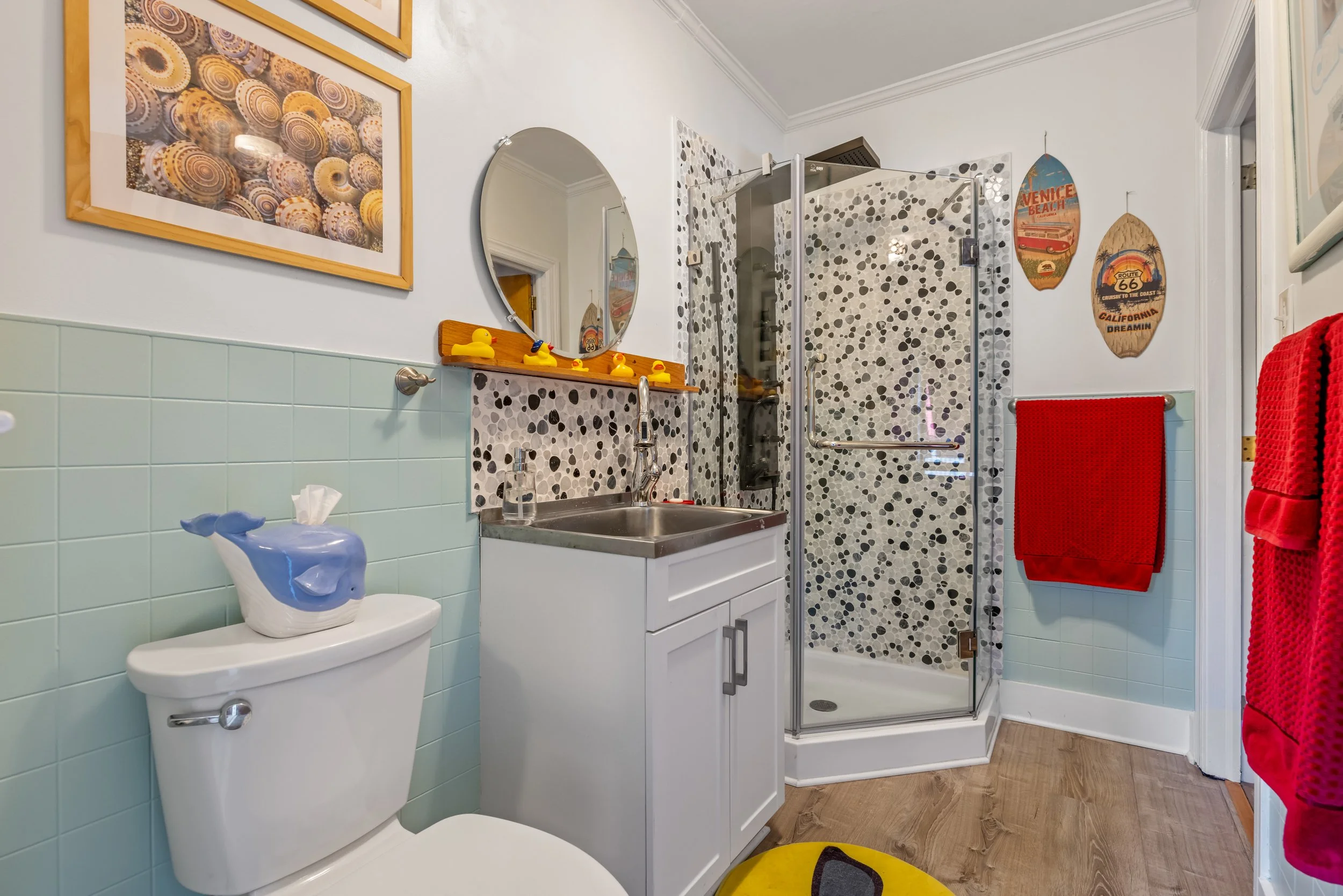 A small bathroom with light green tiling on the lower half of the walls and white paint on the upper half. It features a white toilet with a decorative dolphin tissue holder, a small white vanity with a sink, oval mirror, and rubber duck decor. There