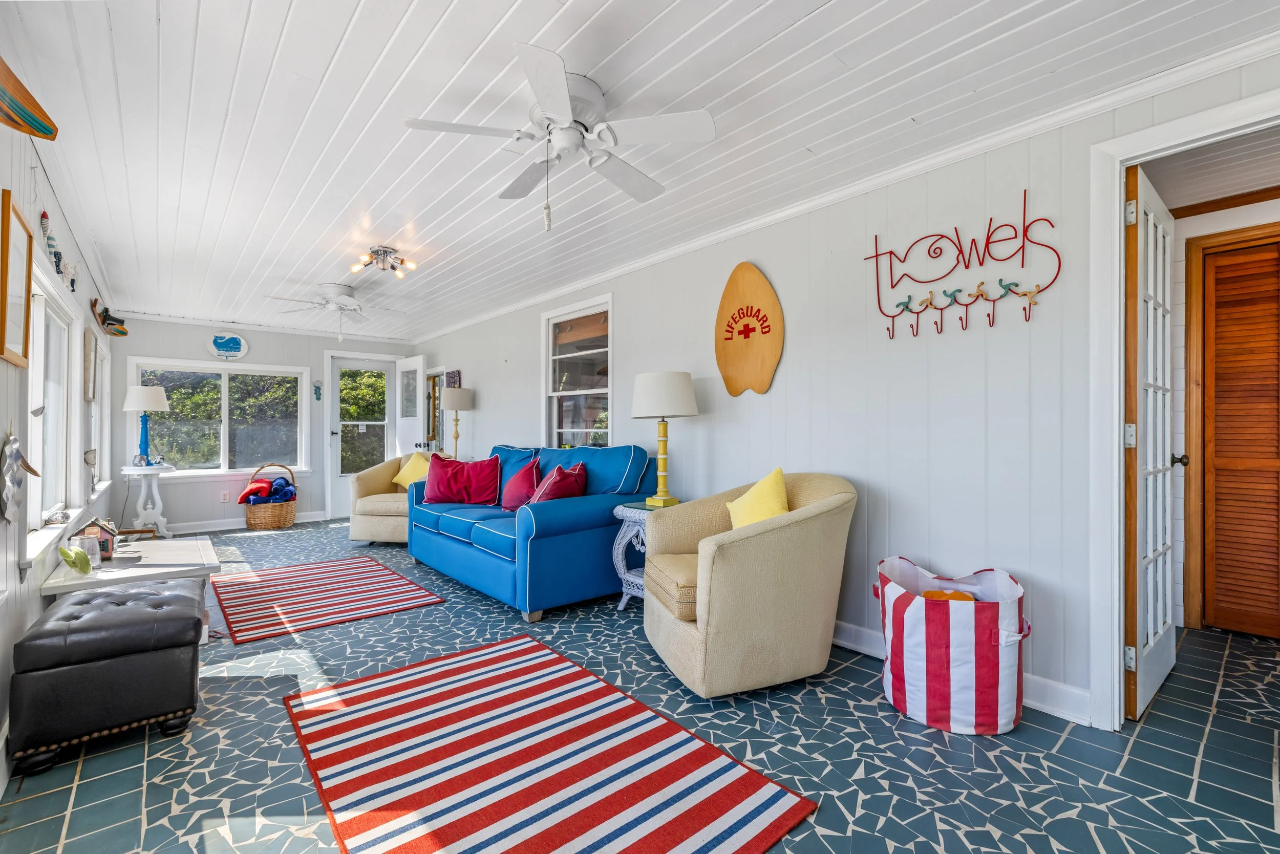 Coastal living room with white shiplap walls and ceiling, ceiling fans, colorful furniture including a blue sofa and beige armchair, striped rugs, and beach decor.