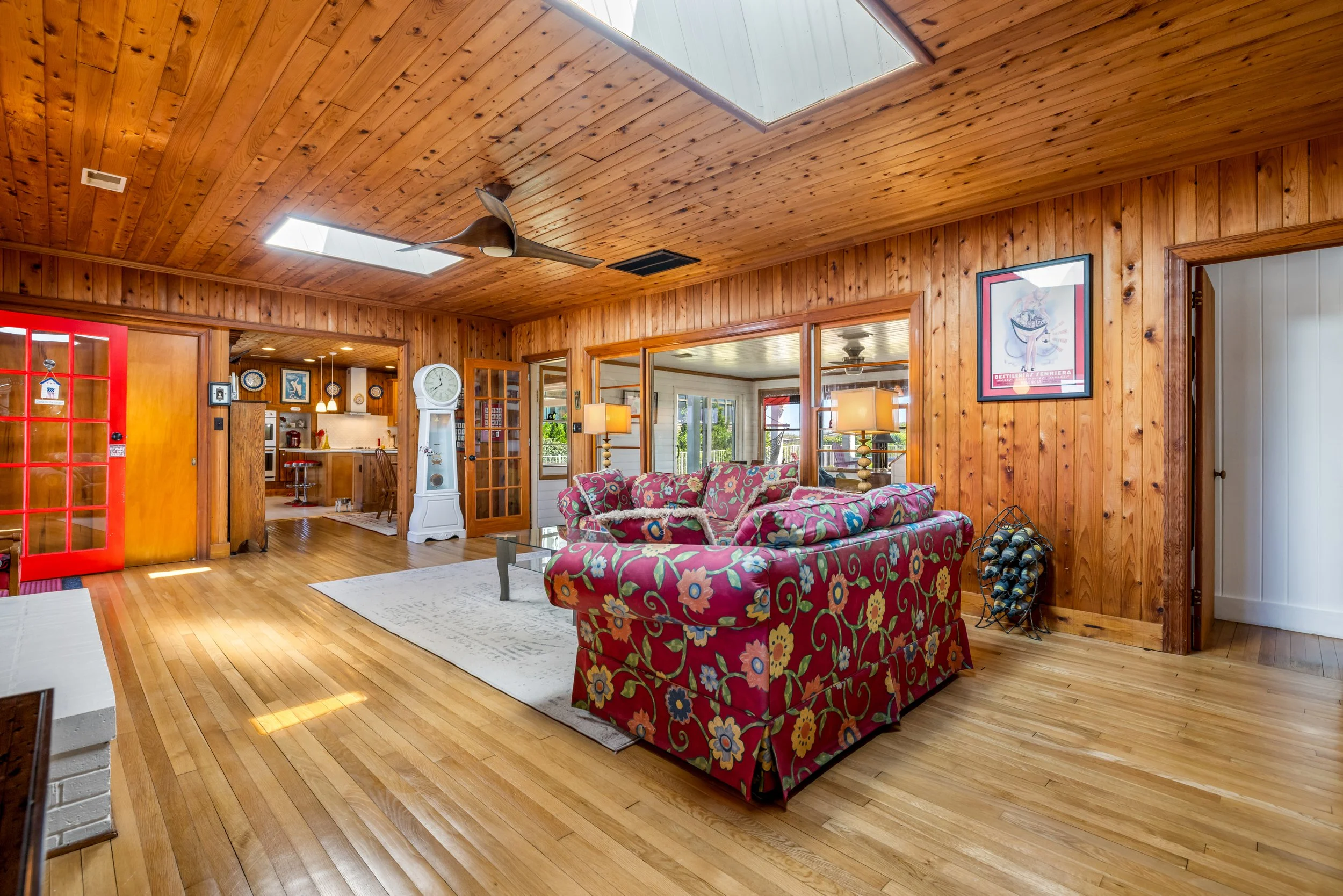 Living room with wooden walls and ceiling, a colorful floral-patterned sofa, a white grandfather clock, and lamps with beige shades. Visible through doorways are a kitchen and dining area.