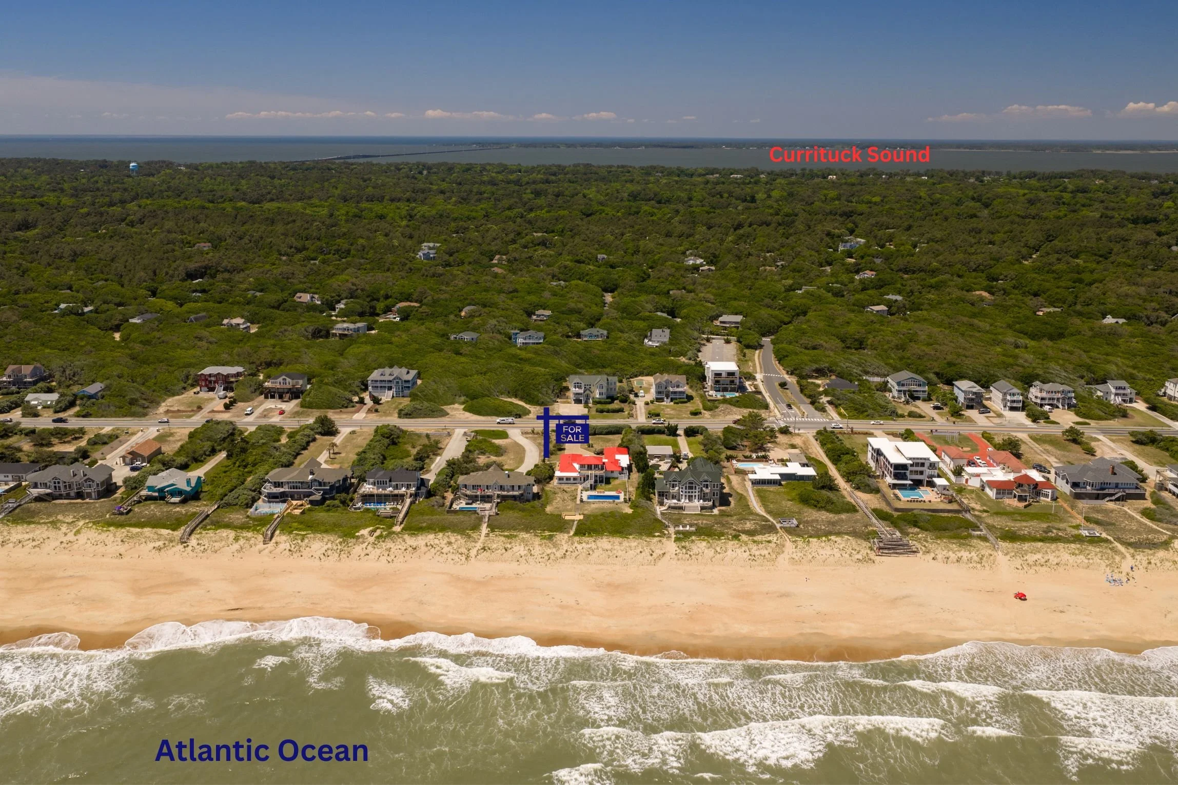 An aerial view of a coastal residential area on a beach with houses, some with pools, and the Atlantic Ocean in the background labeled with red text. The beach and ocean are in the foreground, with waves crashing onto the sandy shore.