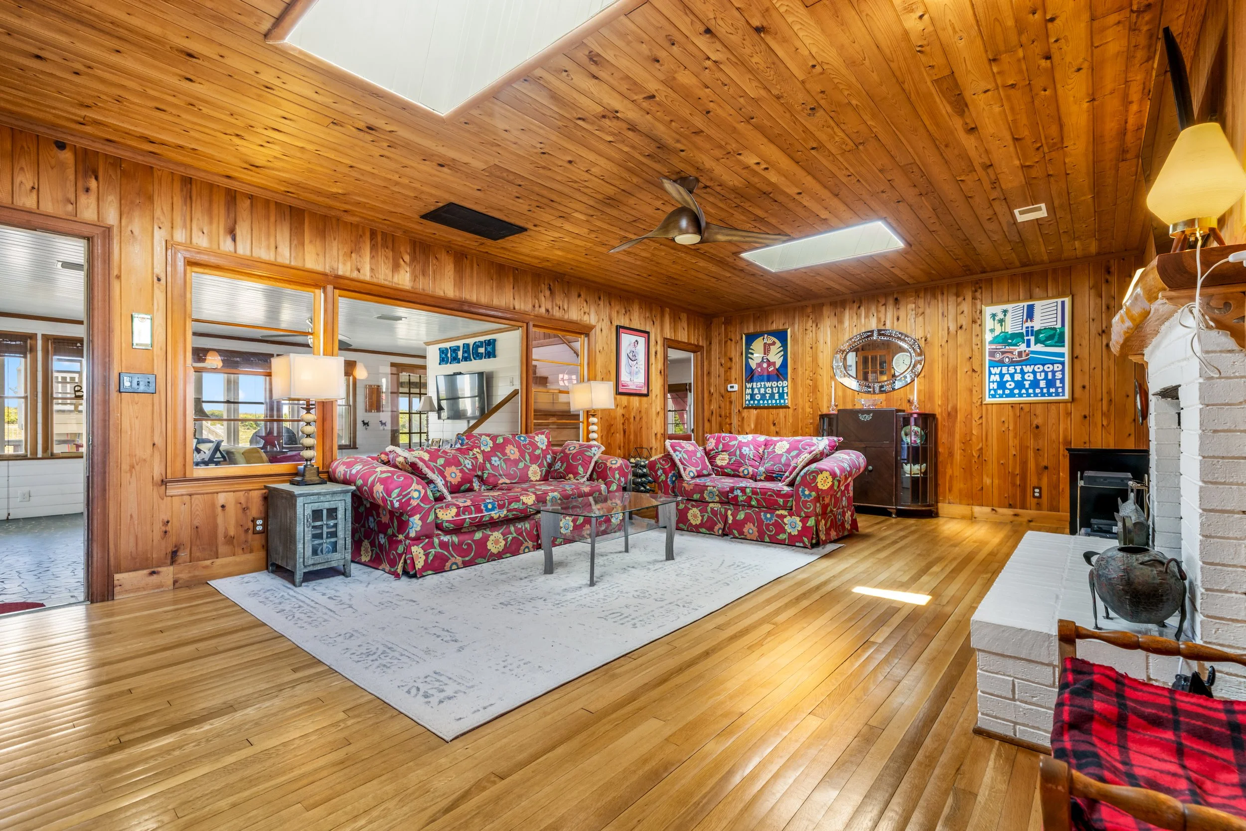 A warmly lit living room with wood-paneled walls and ceiling, featuring floral-patterned sofas, a white rug, and various vintage posters on the walls.