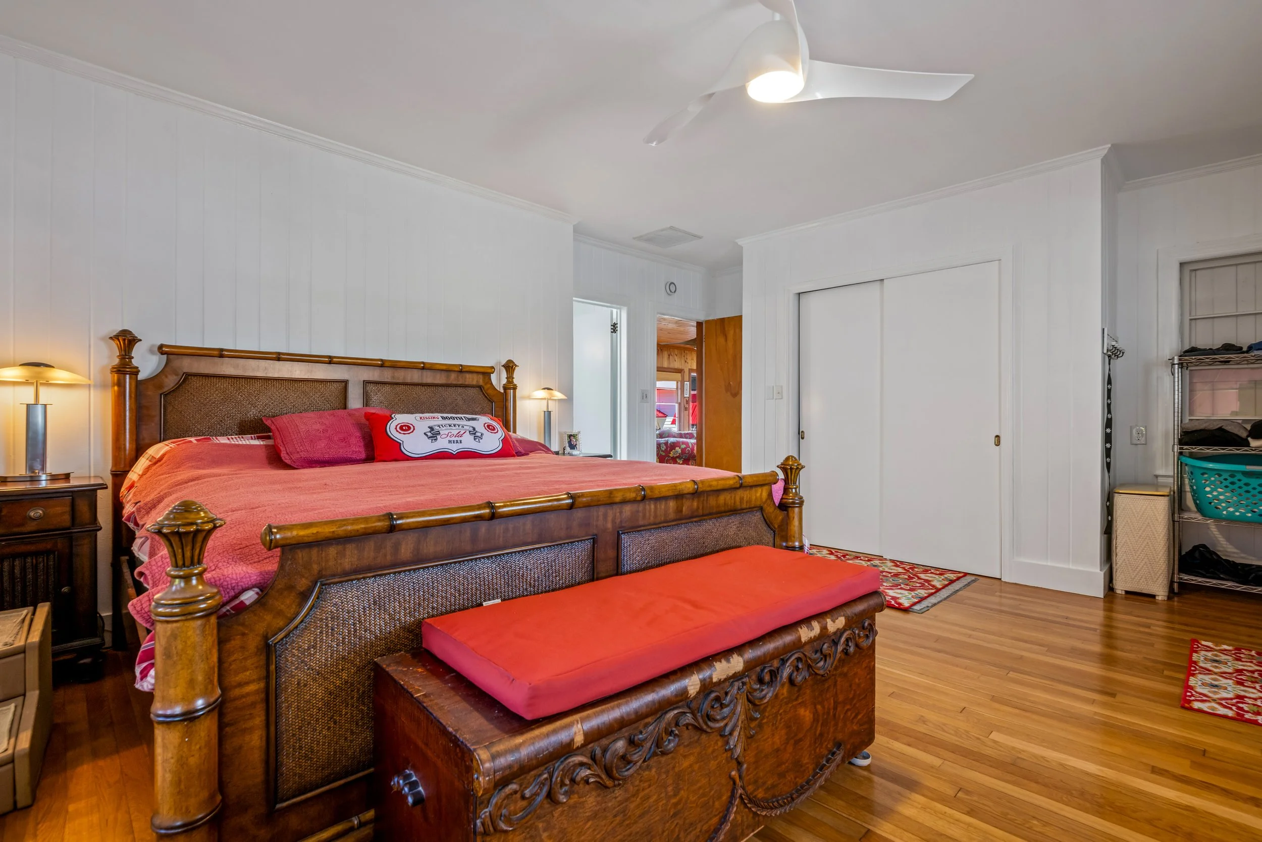 A bedroom with a large wooden bed with an ornate headboard and footboard, pink bedding, and red and pink pillows. There are two lamps on bedside tables, a closet with sliding doors, a shelf with shoes and baskets, and a small area rug on a wooden flo