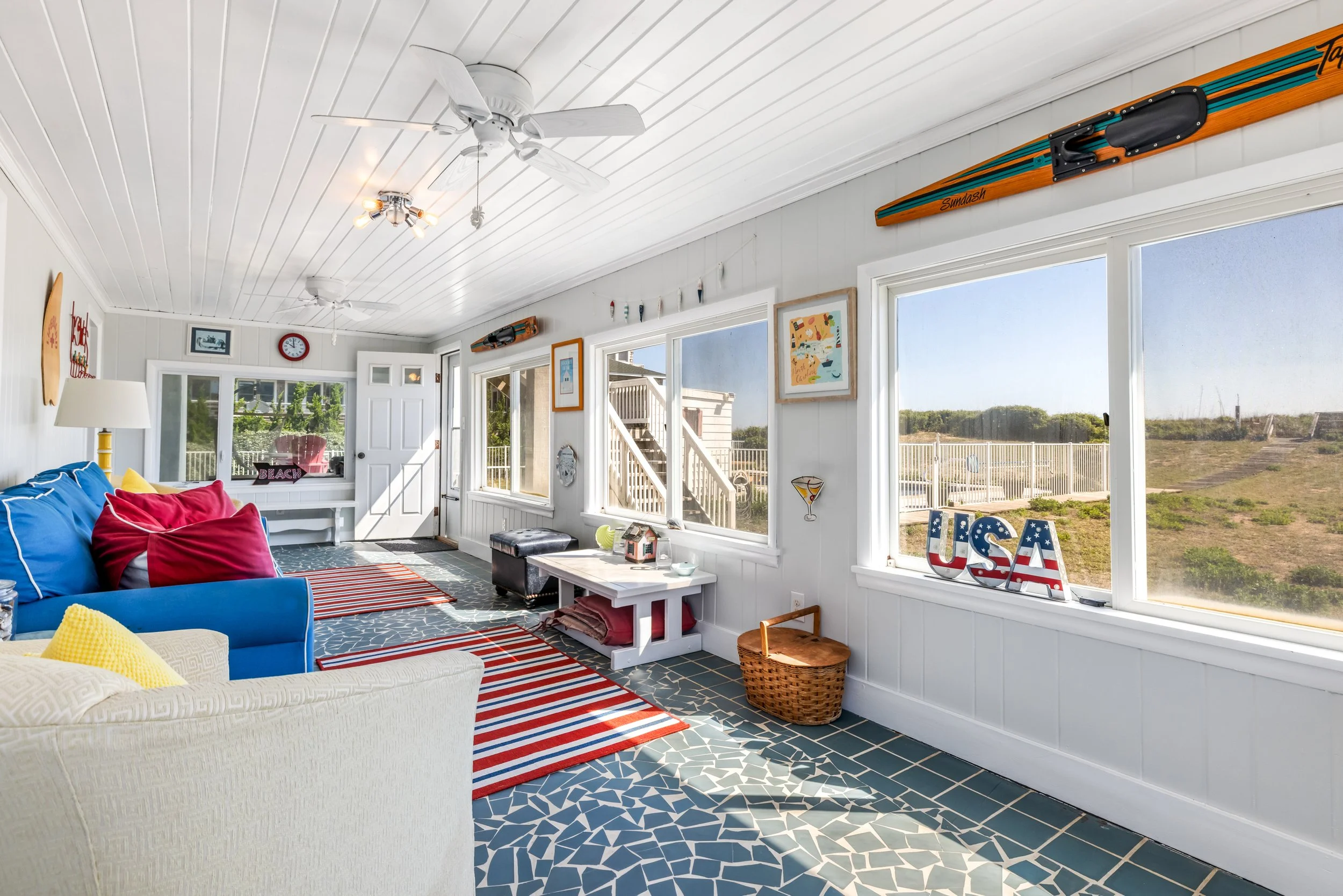 Bright sunroom with white beadboard ceiling, ceiling fans, large windows, and nautical decor including surfboard and USA sign. Colorful cushions on white and blue sofas, patterned rugs, and outdoor view of a backyard with fence.