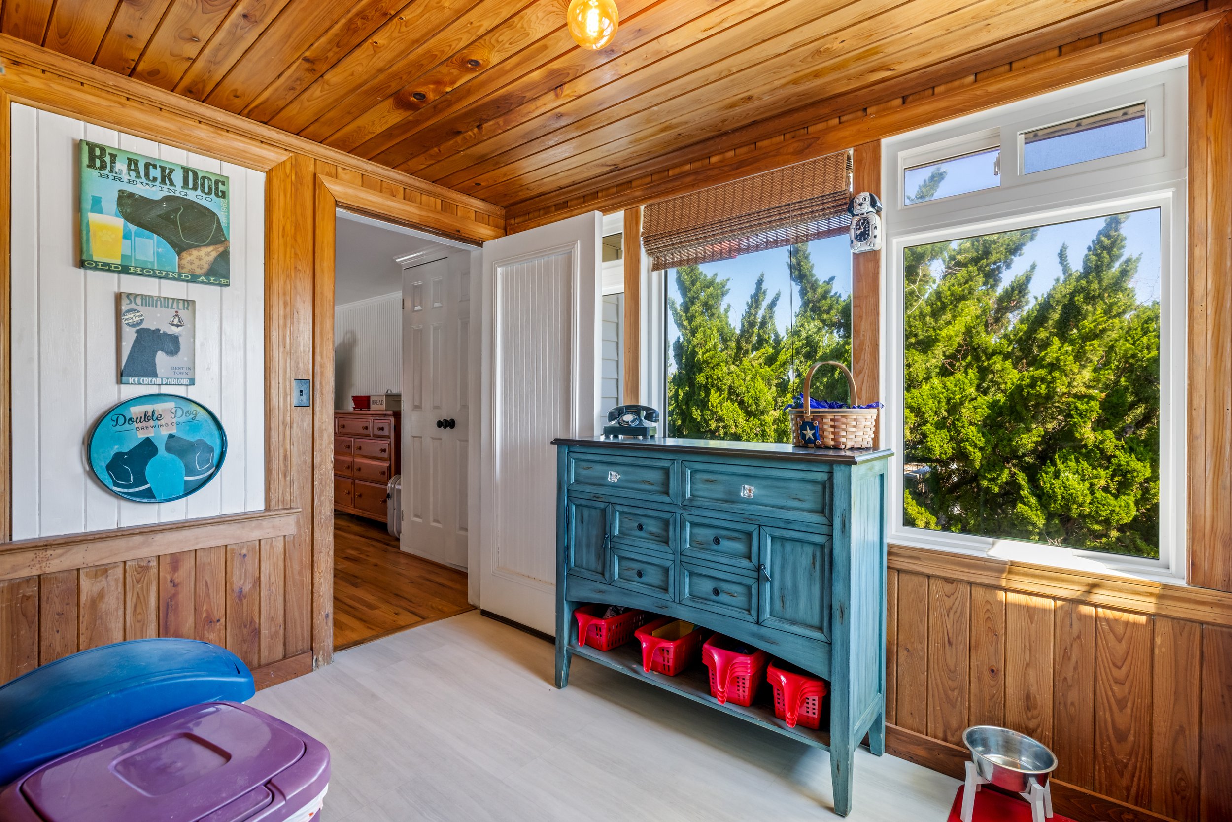 Interior of a room with wood-paneled walls and ceiling, a large window showing green trees outside, a turquoise dresser, colorful bins on a lower shelf, and posters of a dog and a bear on a white wall.