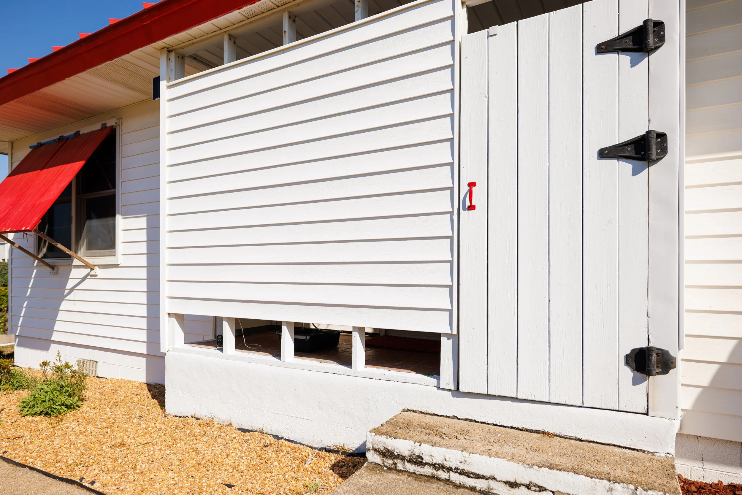 Exterior of a white house with a red awning over a window and a white wooden sliding door on the right side. The house has horizontal siding and is partially built on a raised foundation with some black storage containers underneath. The ground is co