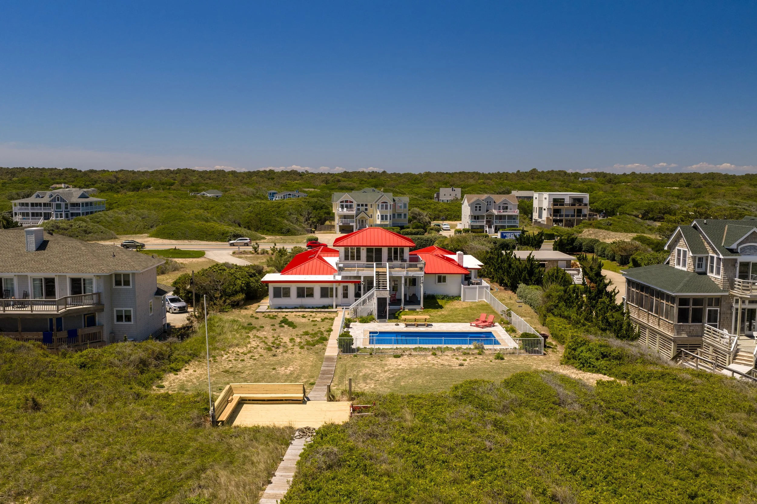 Aerial view of a neighborhood with houses, a swimming pool, and lush green landscape under a clear blue sky.