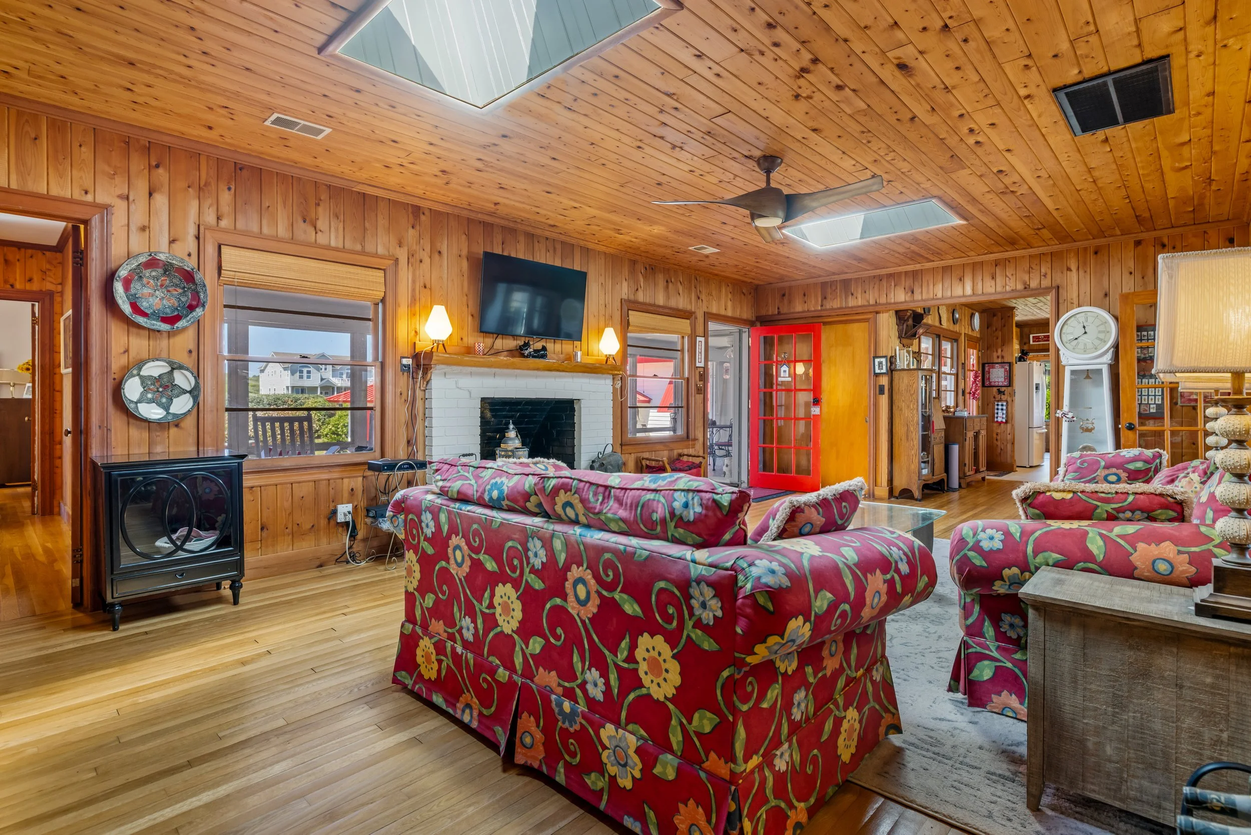 A cozy living room with wood-paneled walls and ceiling, featuring floral-patterned red couches, a television mounted above a white brick fireplace, windows with blinds, and a ceiling fan.