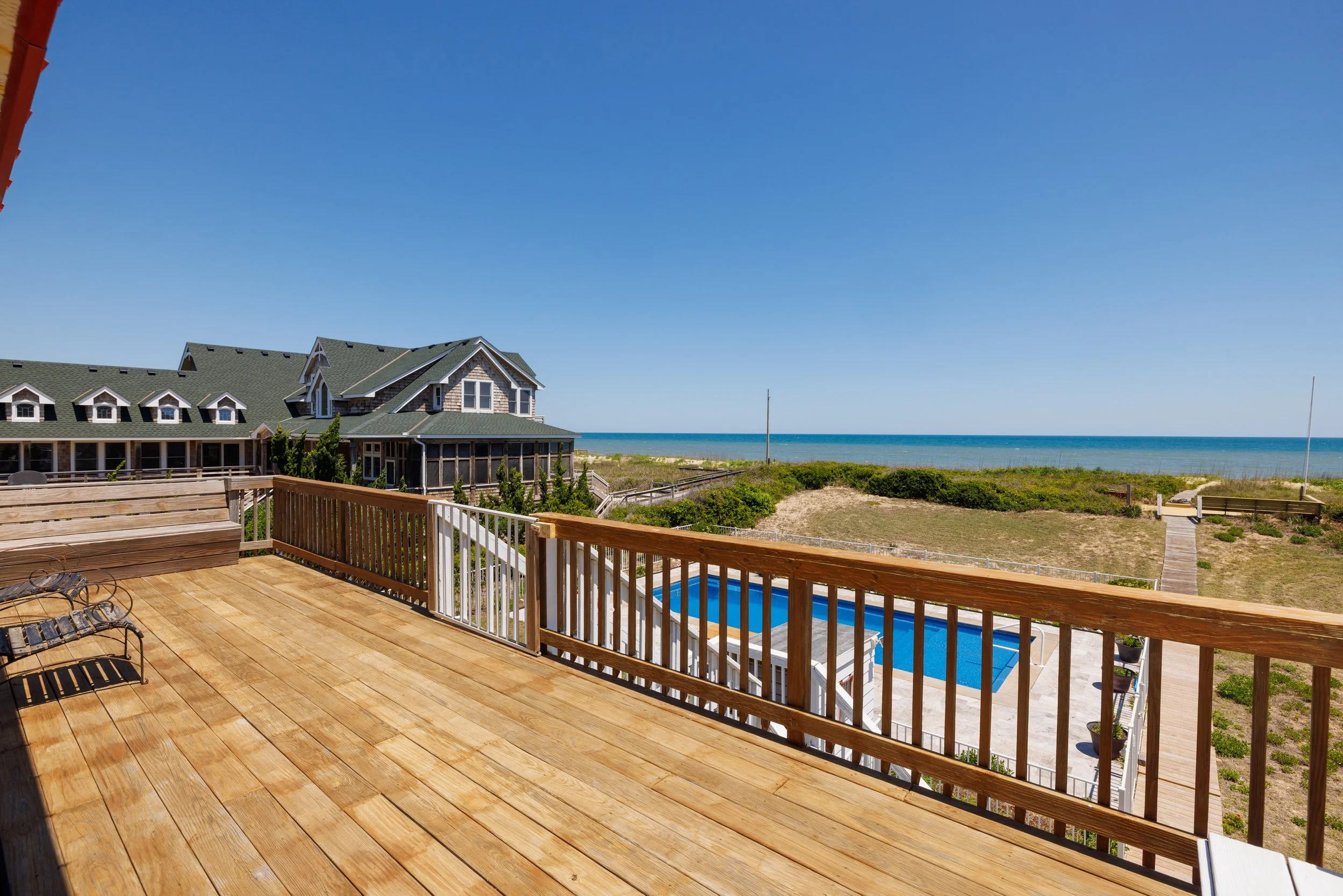 View from a wooden deck overlooking a beach with green shrubbery, a pool below, a house with a gray roof, and the ocean under a clear blue sky.