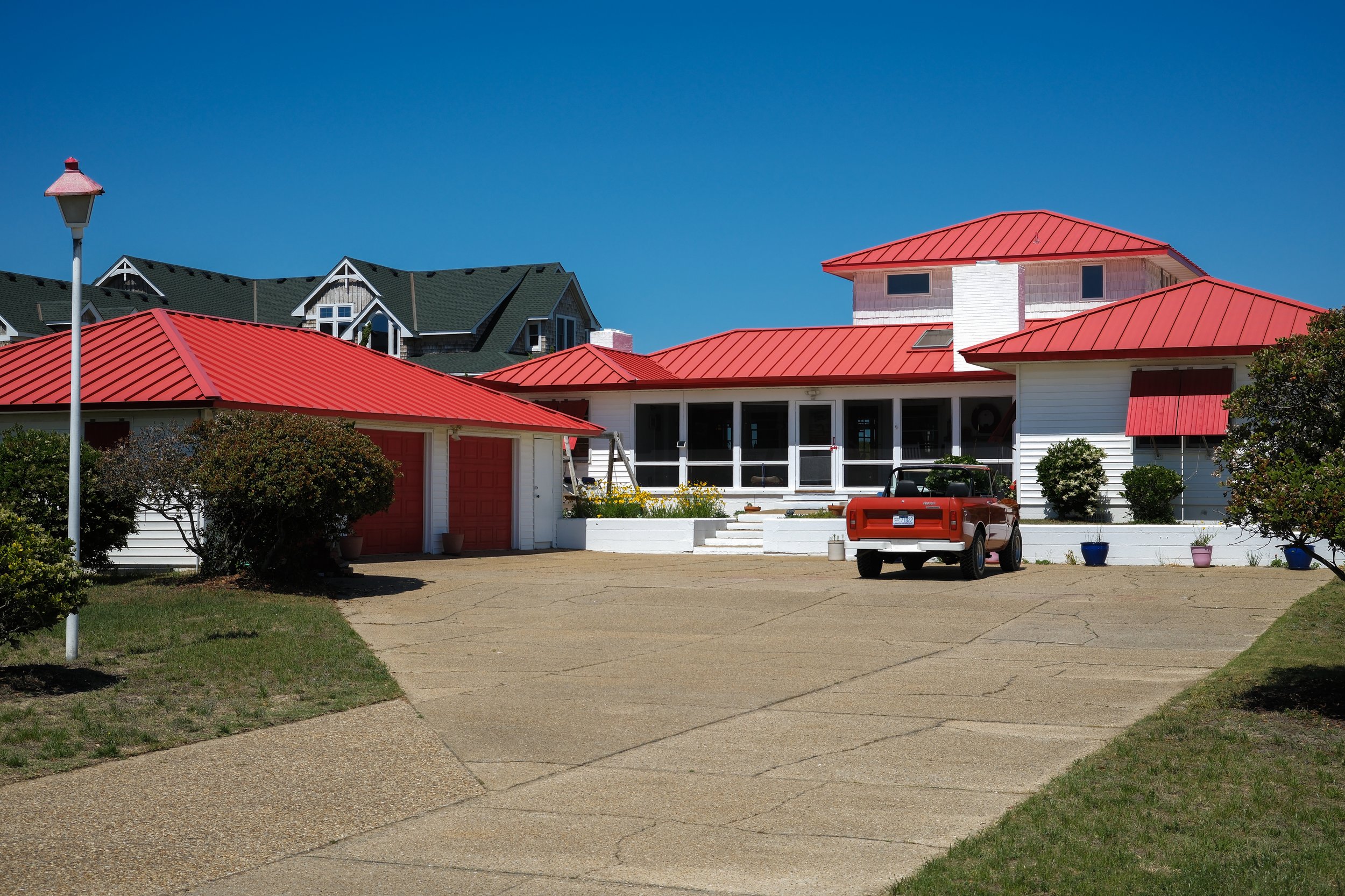 A house with a red metal roof, white walls, large porch, and multiple windows. There is a driveway with a red pickup truck and potted plants along the edge. A black golf cart is parked near the house, and a tall street lamp is visible on the left. Th