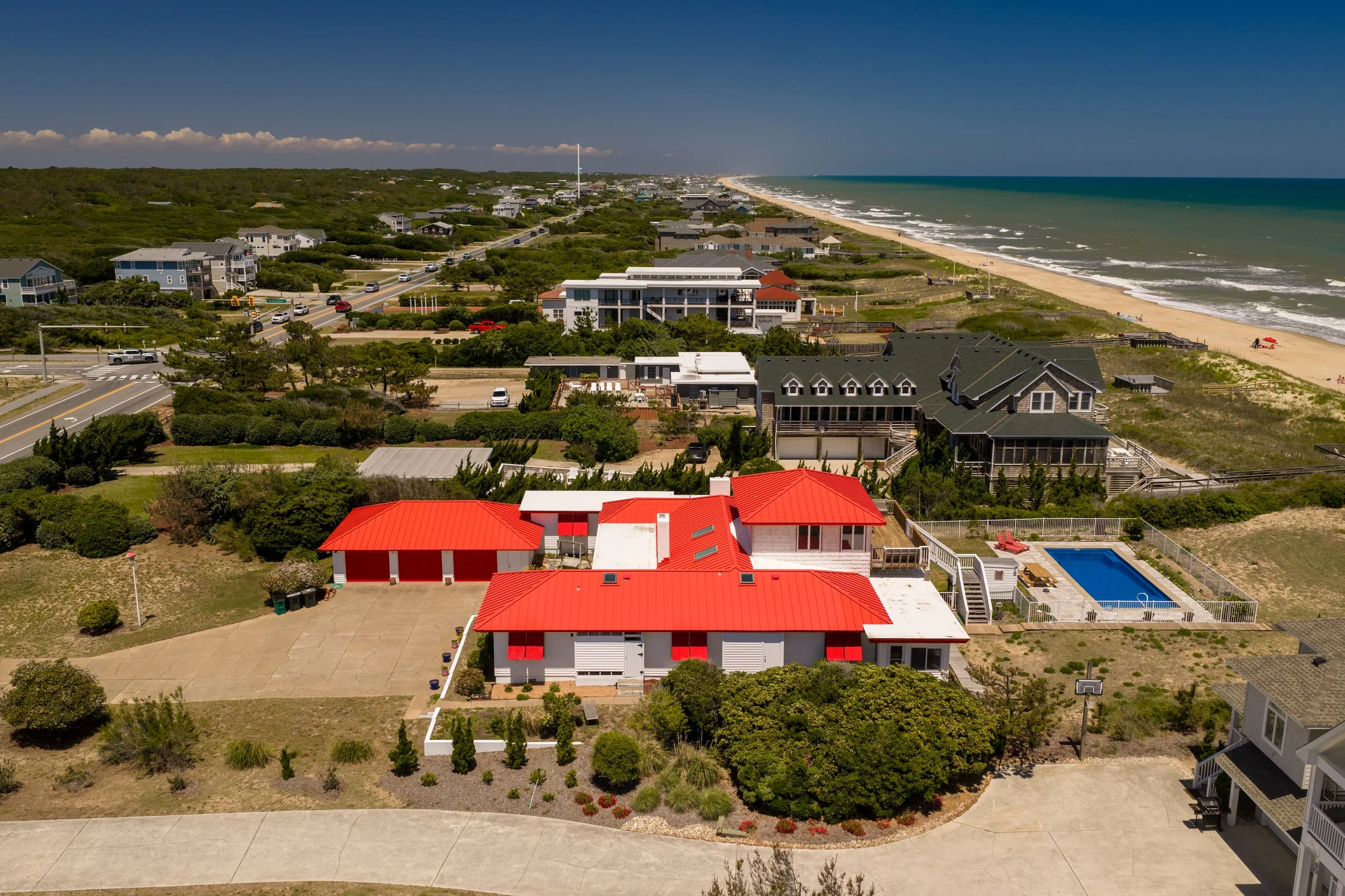 Aerial view of a coastal neighborhood with houses, trees, and a beach along the ocean, featuring a blue pool and a red-roofed building in the foreground.