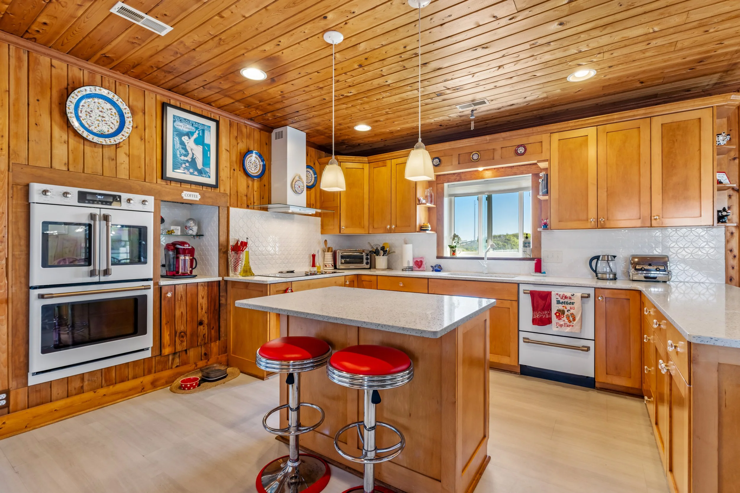 A kitchen with wooden cabinets and ceiling, white countertops, a white oven, a toaster, and a window. Two red barstools are in front of an island in the center.